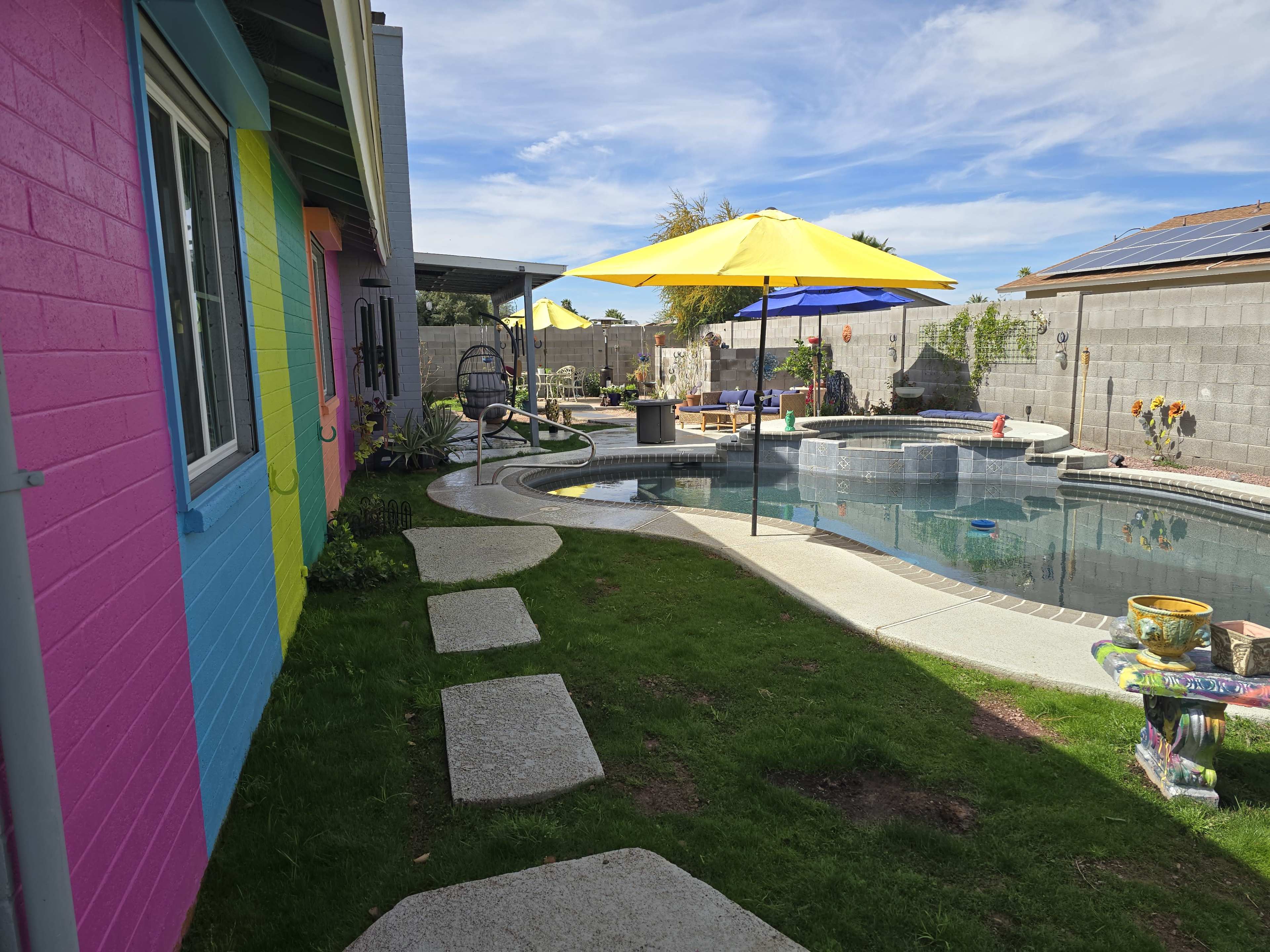 A colorful house with a pink side wall is adjacent to a swimming pool area featuring yellow umbrellas and a stone pathway.