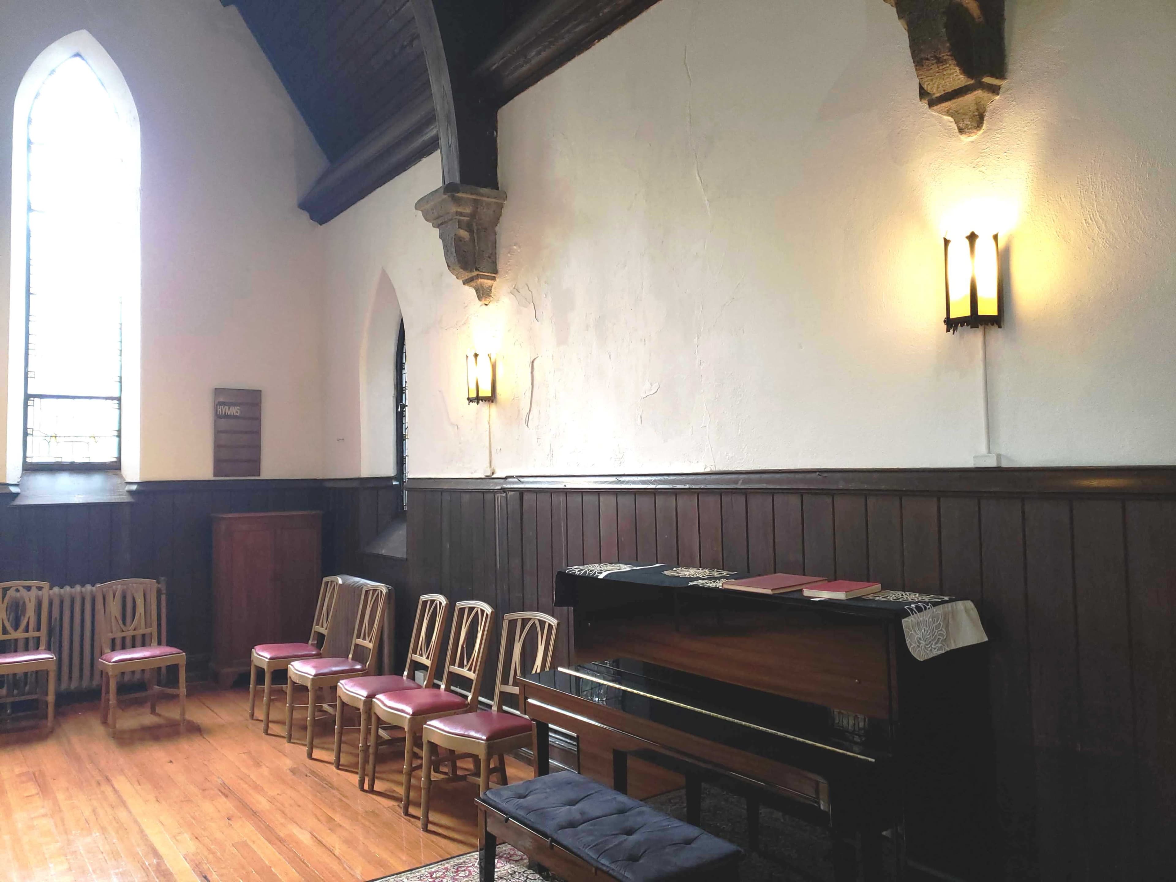 A small, empty room with wooden flooring, a piano against one wall, and several chairs arranged in front of it.