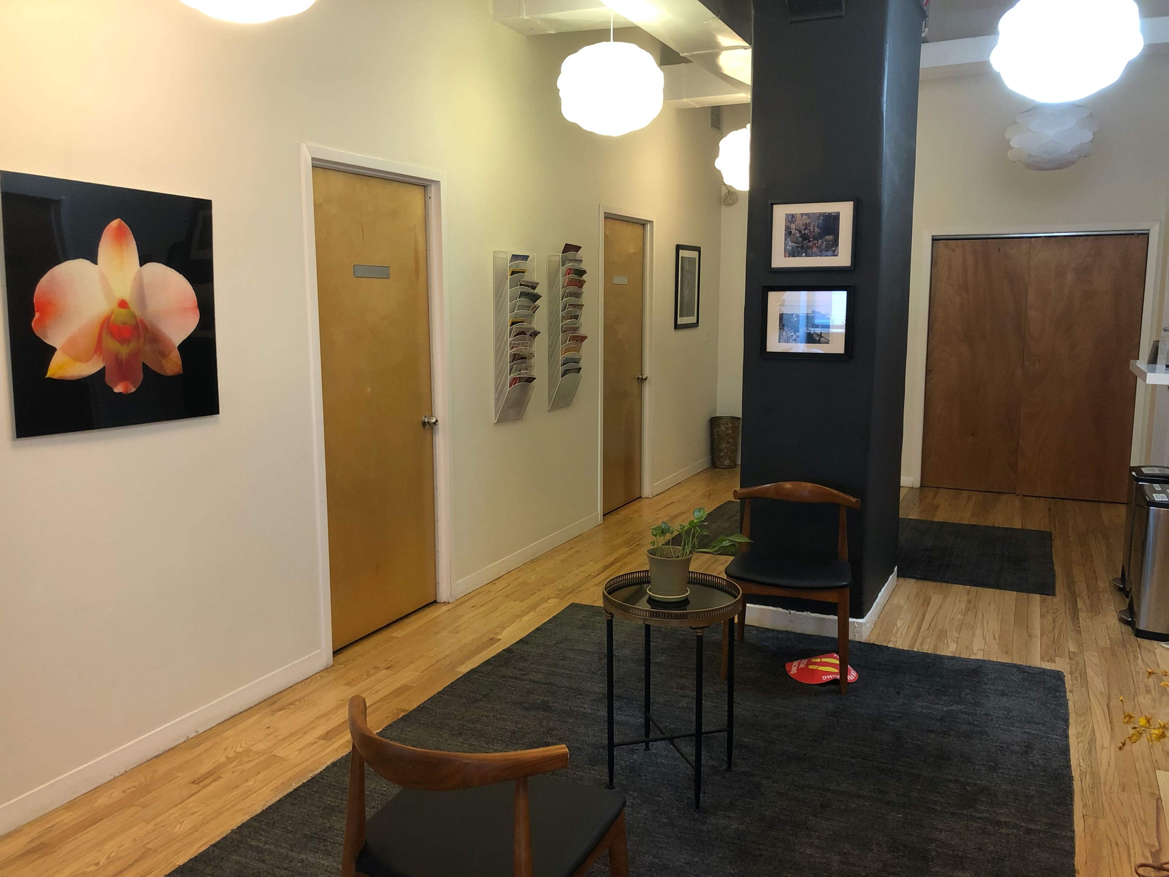 A corridor with beige walls, two wooden doors on the left, a gallery wall with framed pictures on the right, and two chairs beside a small table in the center, illuminated by ceiling lights.