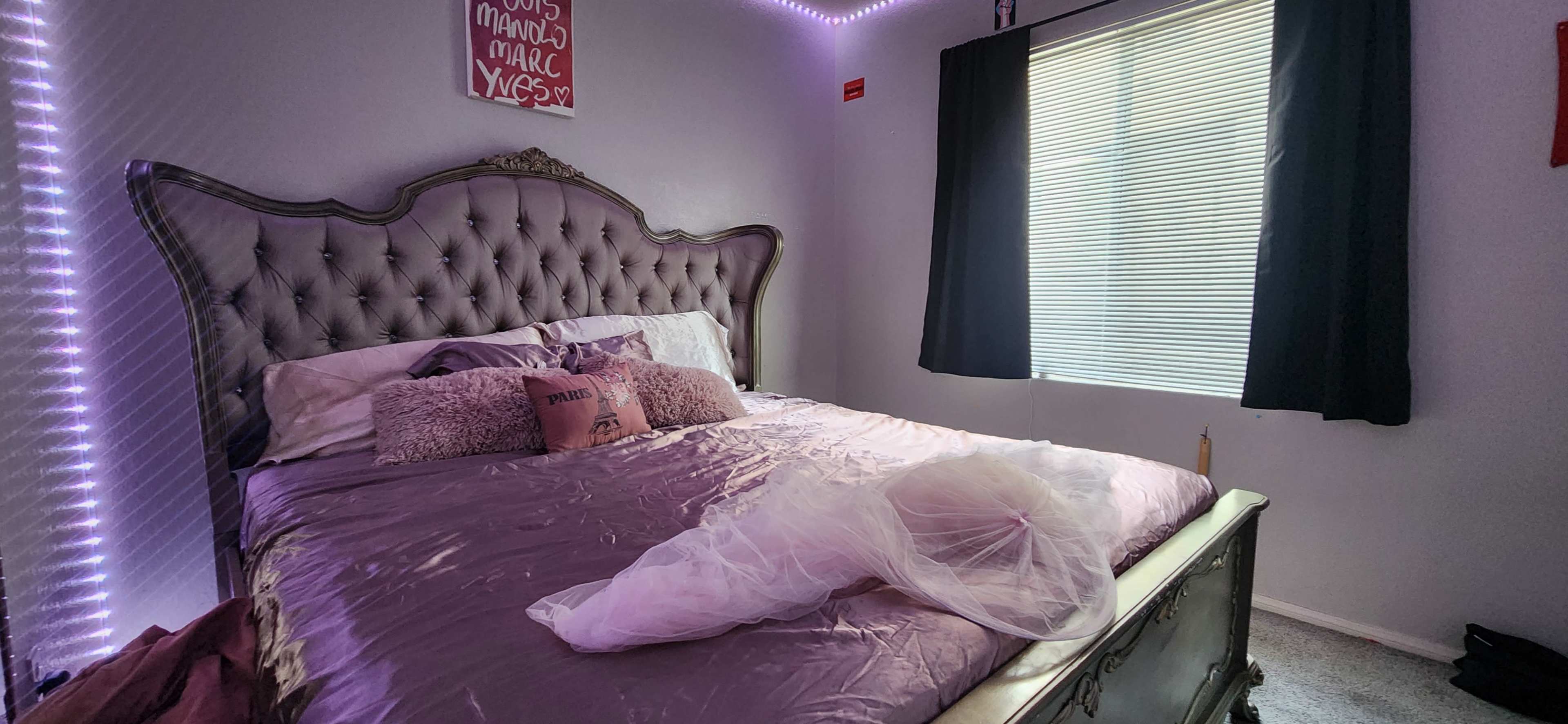 A bedroom features a large, tufted headboard bed with purple bedding and a window covered by black curtains.
