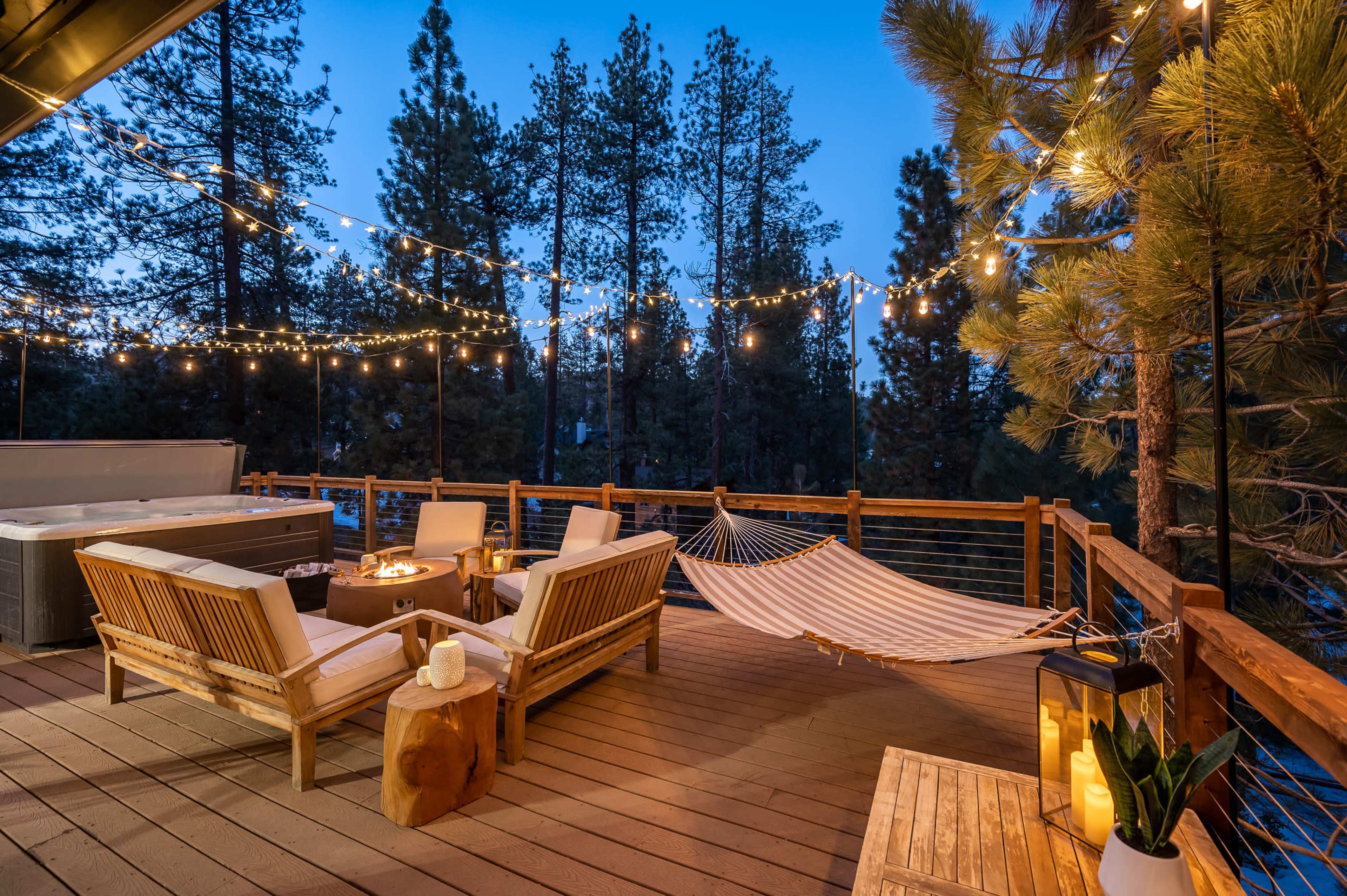A deck with wooden seating, a hammock, and string lights is surrounded by tall pine trees at dusk.