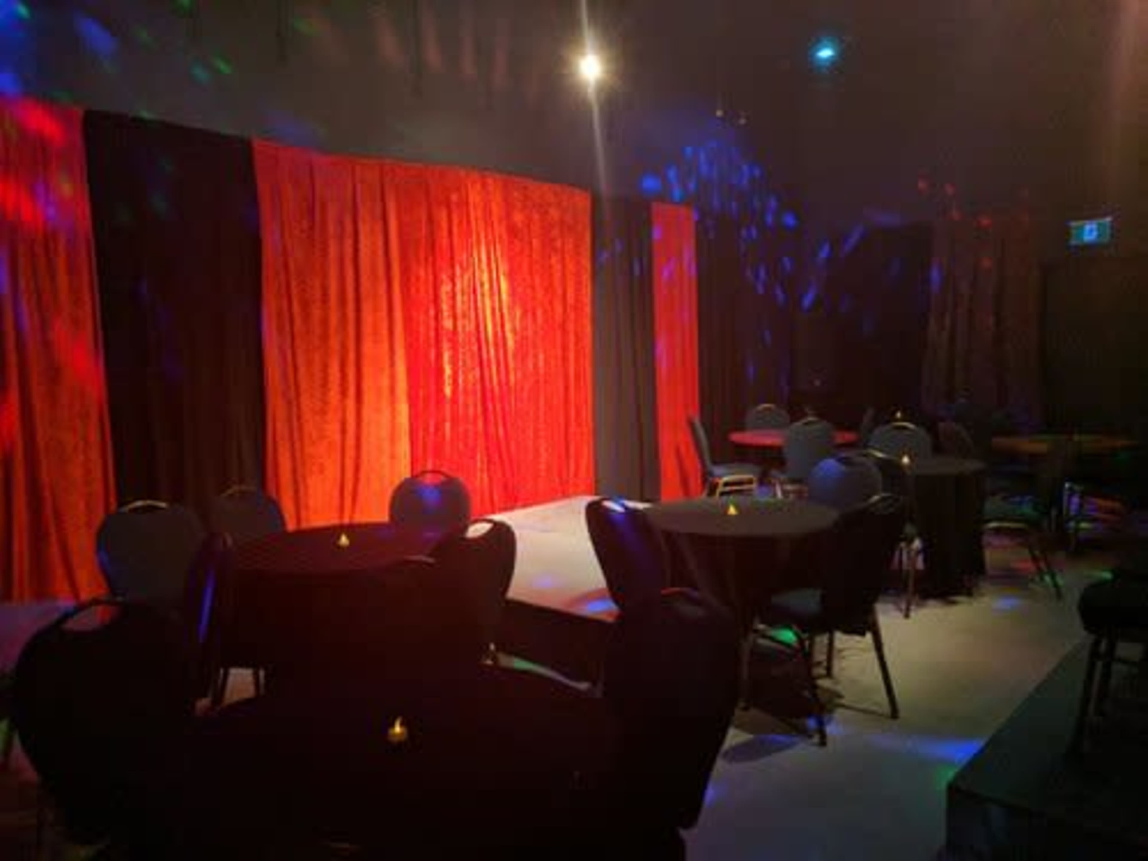 A dimly lit stage adorned with red and black curtains in a room with circular tables and chairs arranged for an audience.