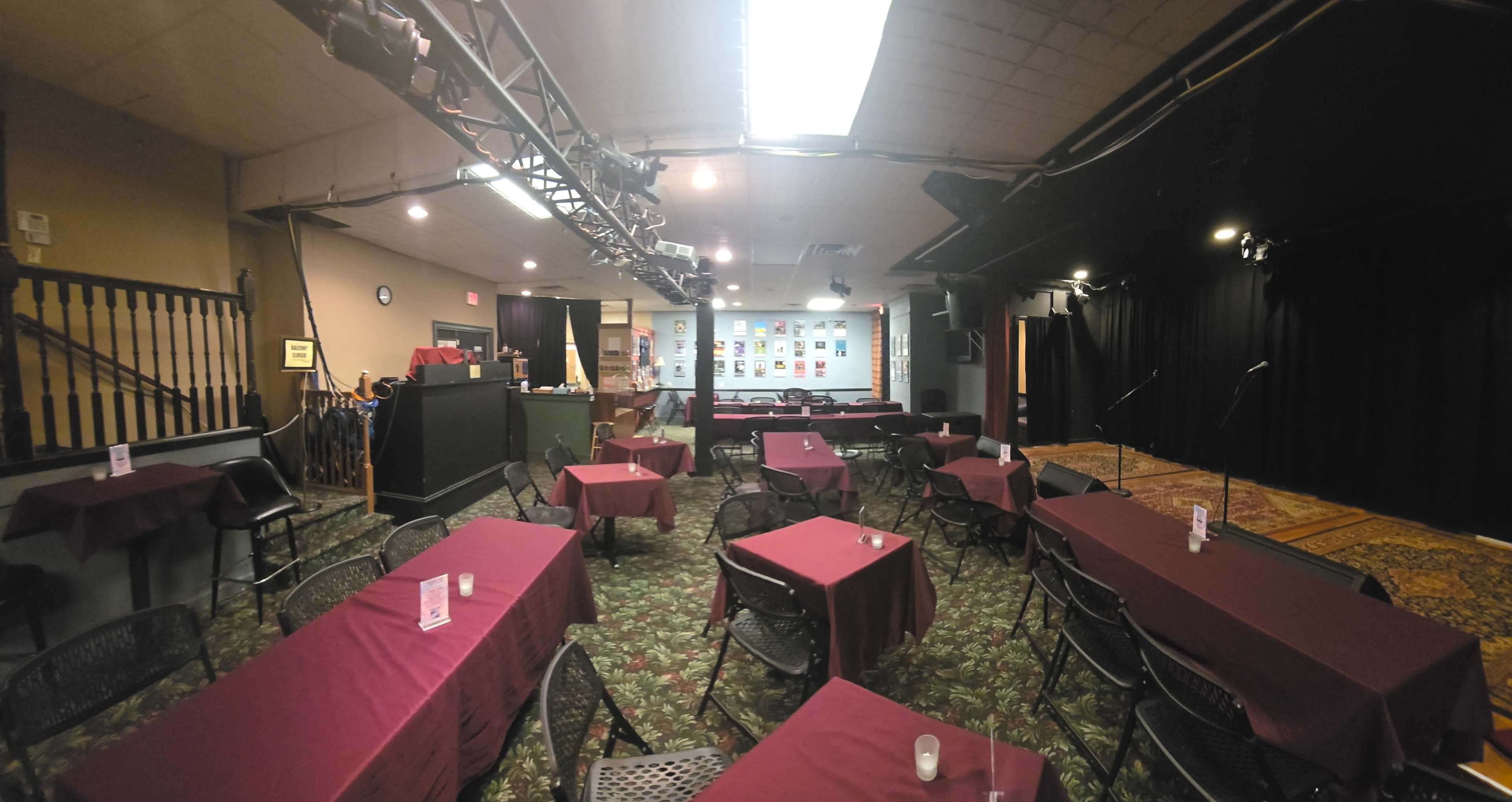 The image shows a performance venue set up with maroon tablecloth-covered tables arranged in a semi-circle facing a small stage with a backdrop of framed posters.