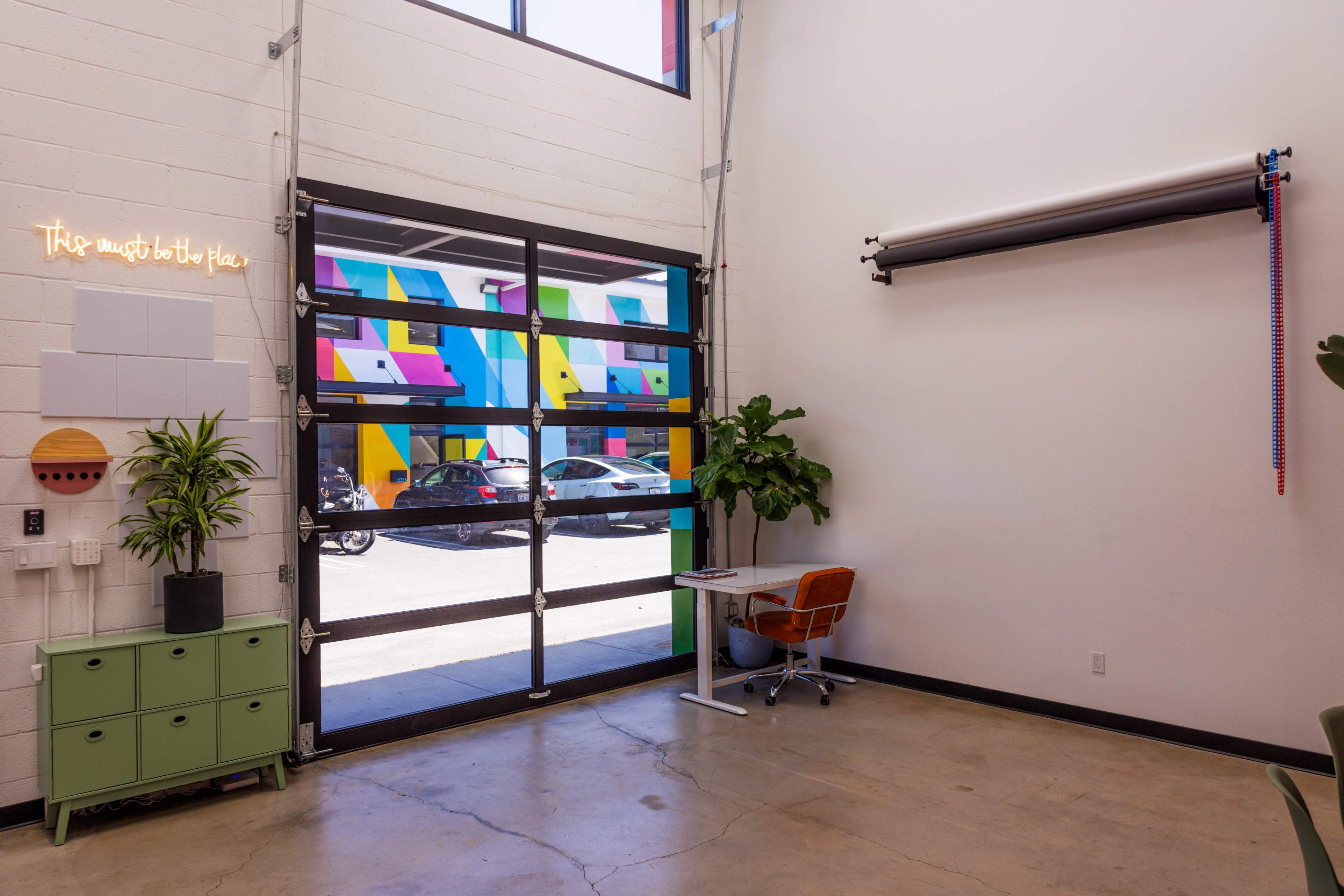 The image shows an interior space with a large open garage door revealing colorful murals outside, a small desk and chair, a green cabinet, and a potted plant.