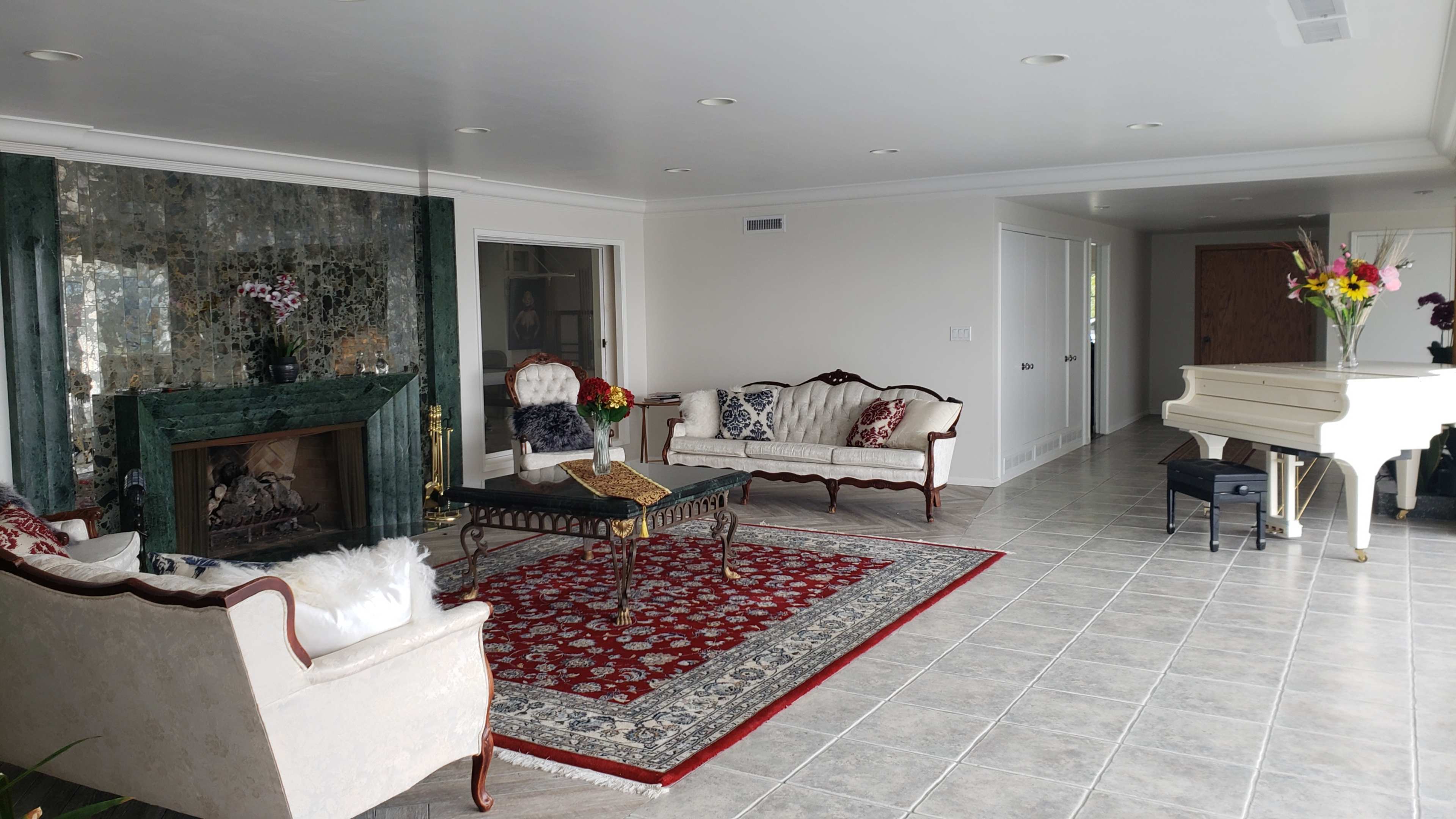 The spacious living room features a green marble fireplace, two elegant sofas, a coffee table on a patterned rug, and a grand piano near a wall of windows.