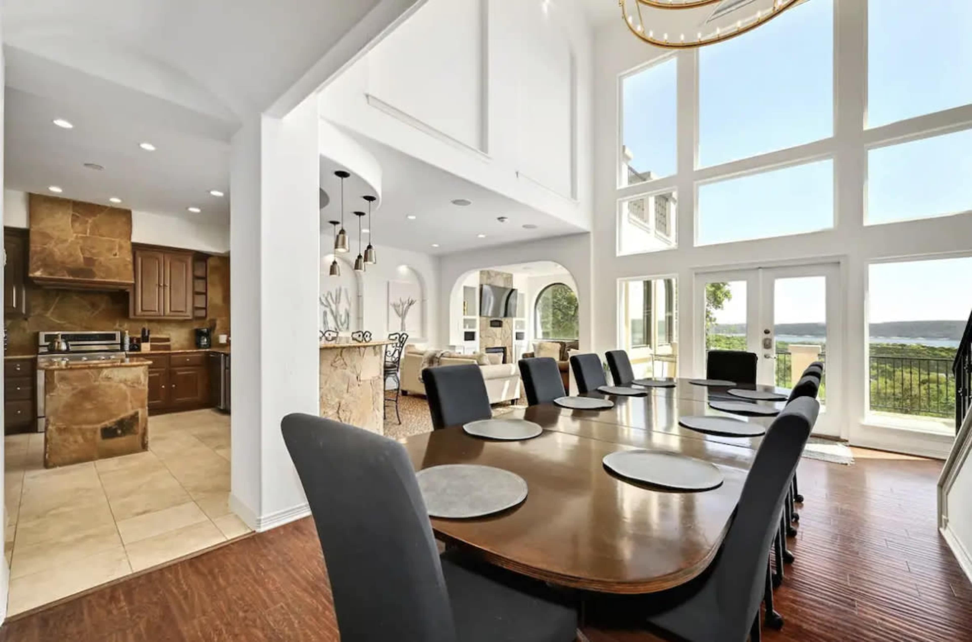 A spacious dining area with a large wooden table and black chairs, adjacent to a modern kitchen with stone accents and large windows overlooking a scenic view.