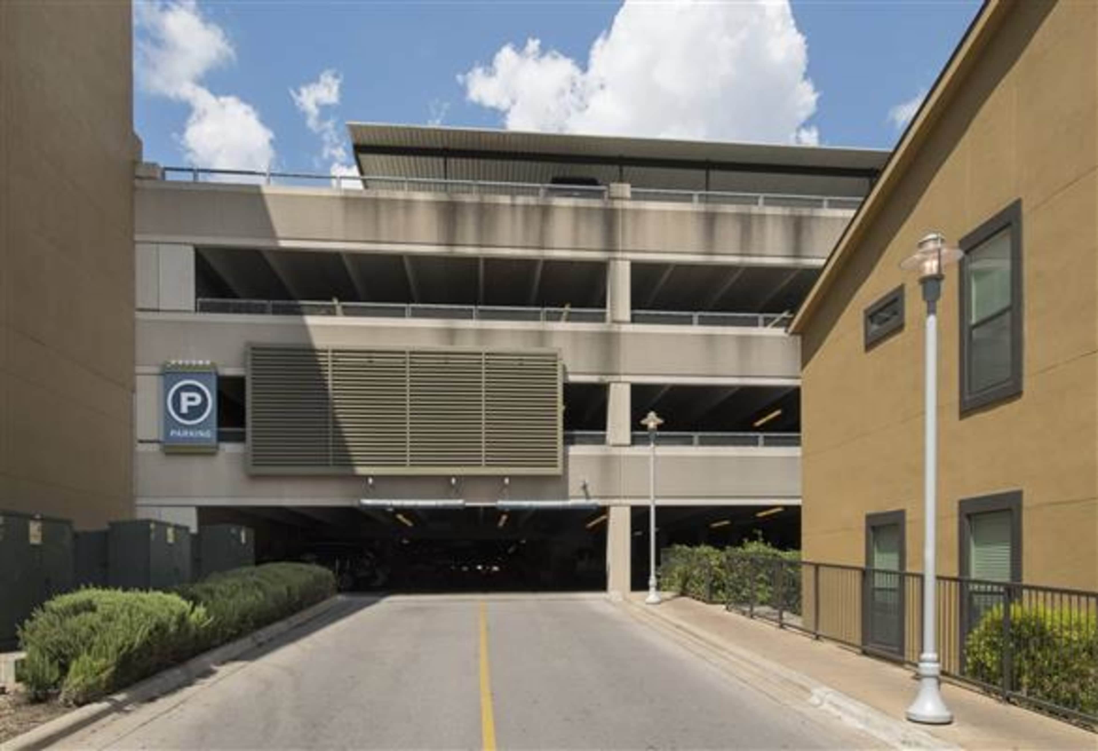 A multi-level parking garage is seen at the end of a driveway, flanked by buildings and landscaping on either side.