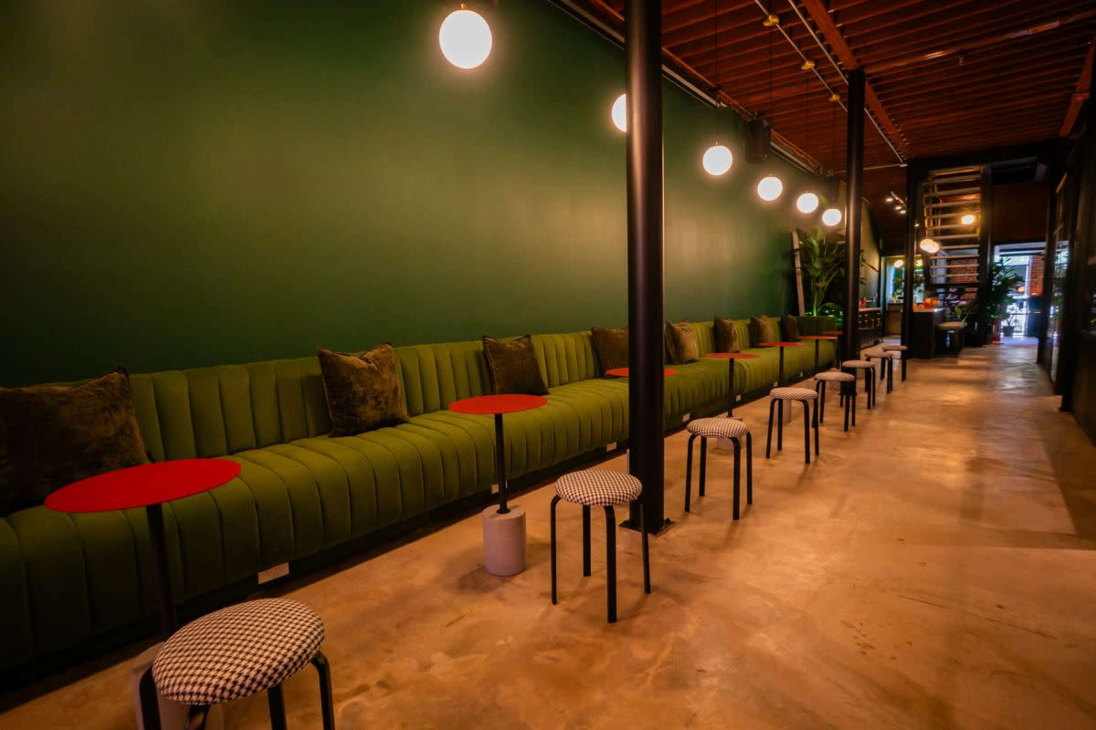 The image shows a modern interior of a cafe or lounge with a green wall, long green seating, small red tables, and light fixtures overhead.