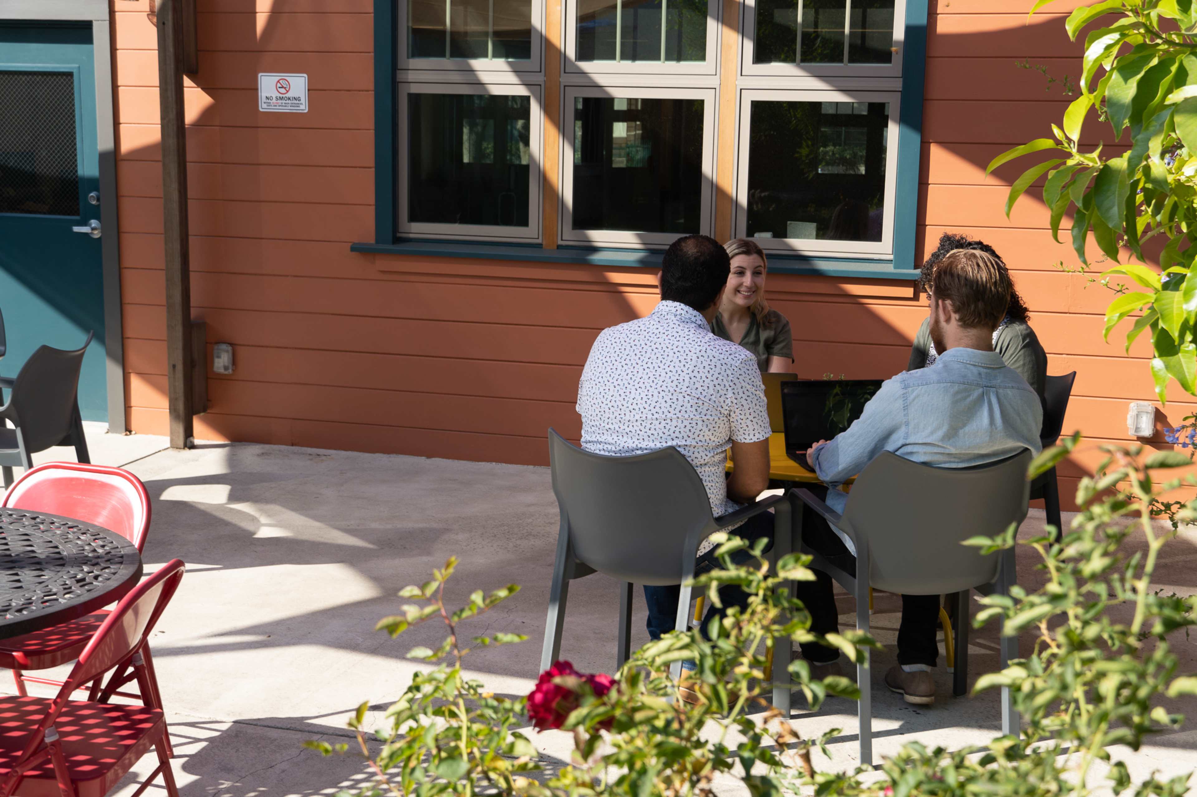 Four people sit at a table in an outdoor setting, engaged in conversation near a building with large windows.