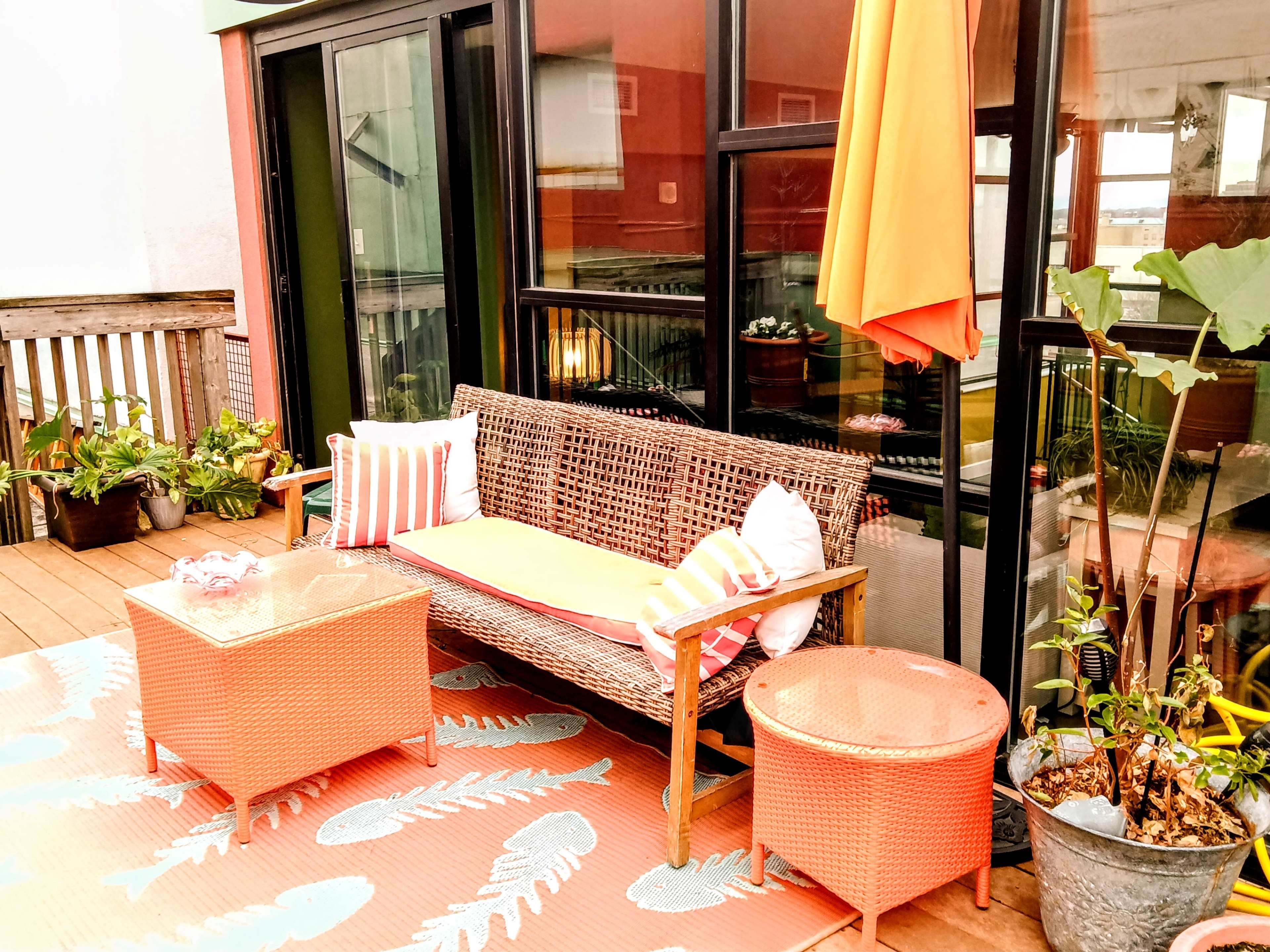 A patio with a wicker sofa, orange cushions, a glass coffee table, and potted plants, set against a backdrop of large glass doors.