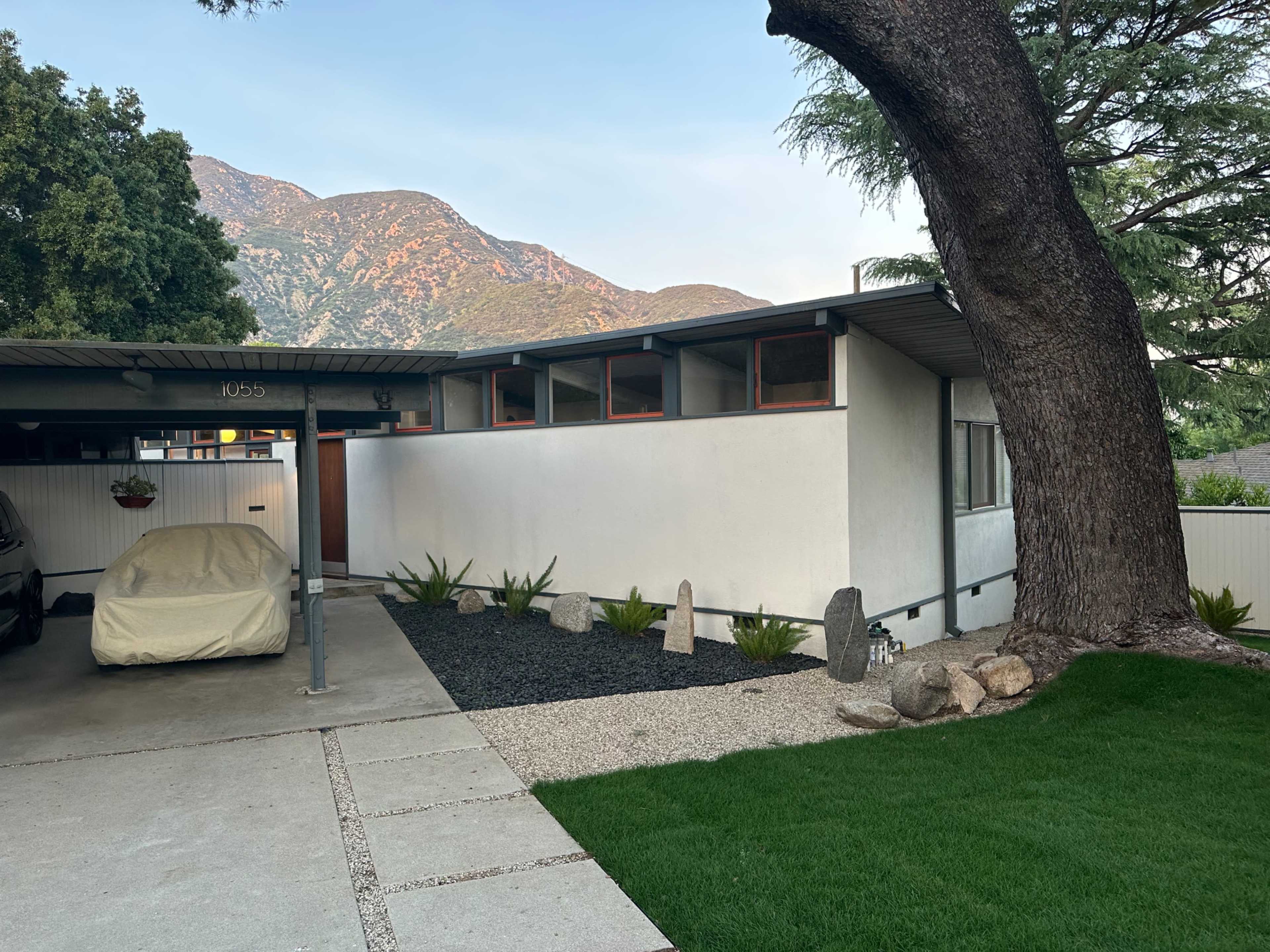 A modern house with a carport and a covered vehicle is situated next to a gravel and grass landscape, with mountains in the background.