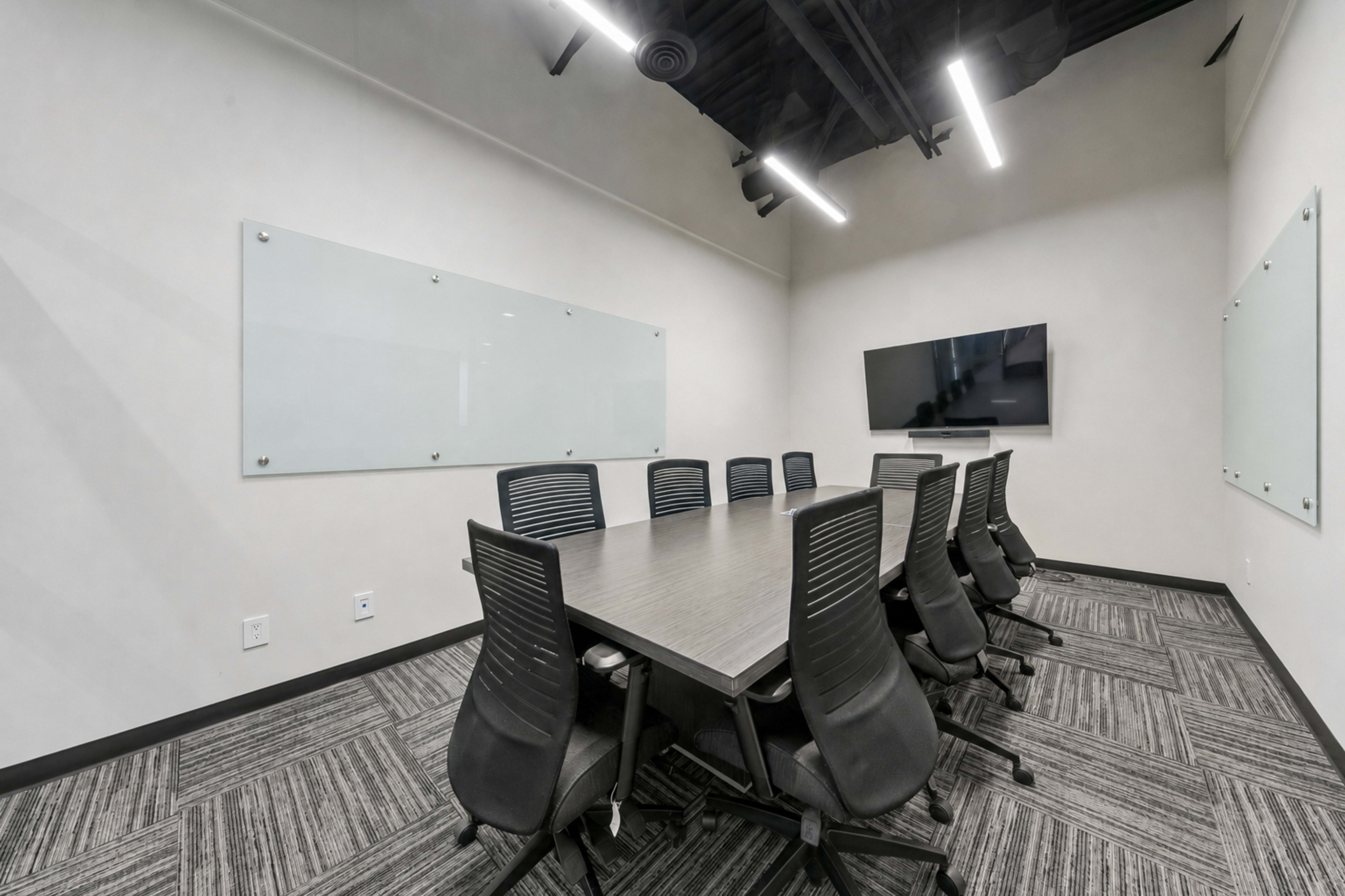 A modern conference room features a long table surrounded by ergonomic chairs, with presentation screens and glass boards on the walls.
