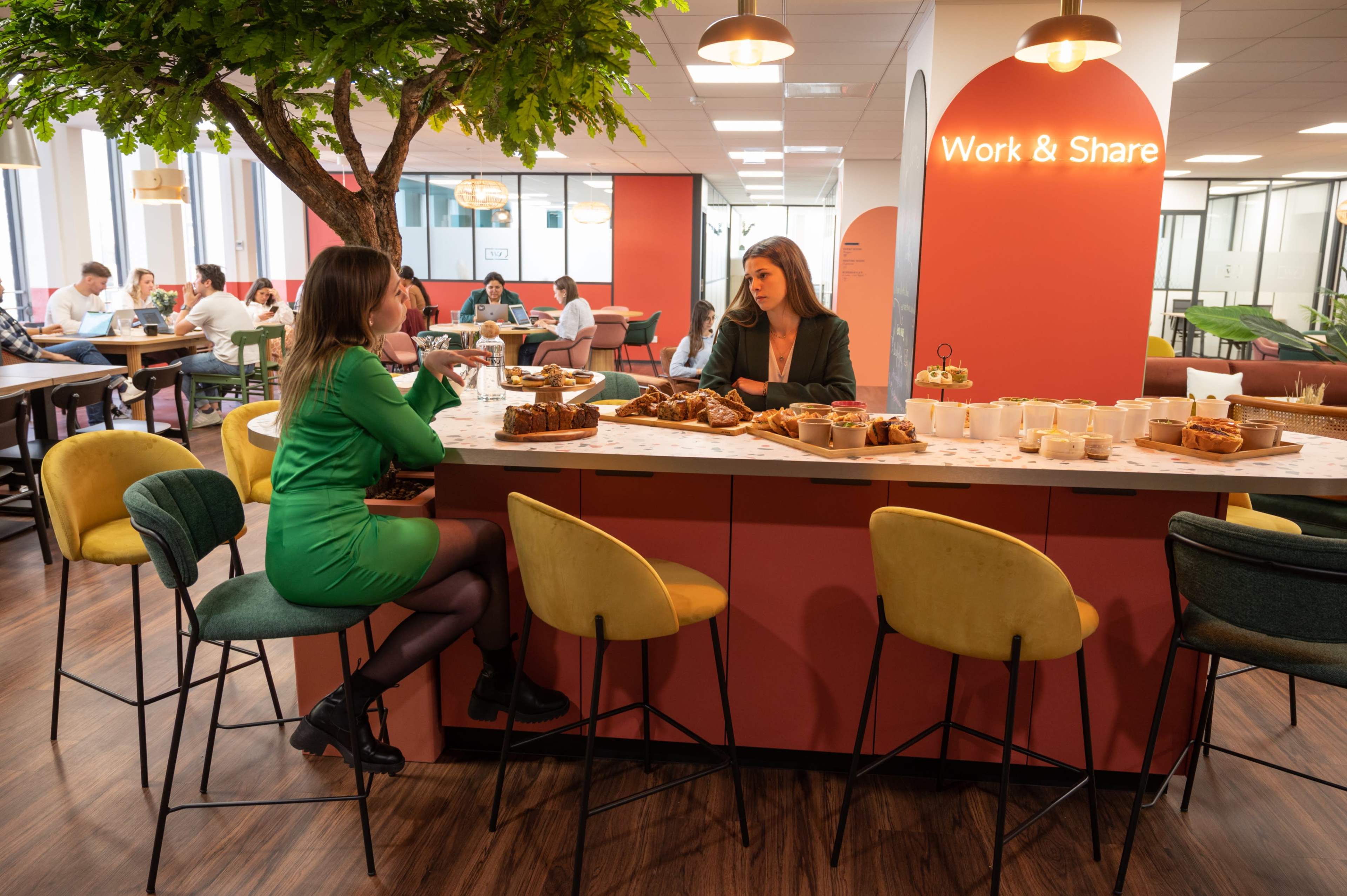 Two women sit at a bar-style table in a modern office space with pastries on display, while other people work in the background.