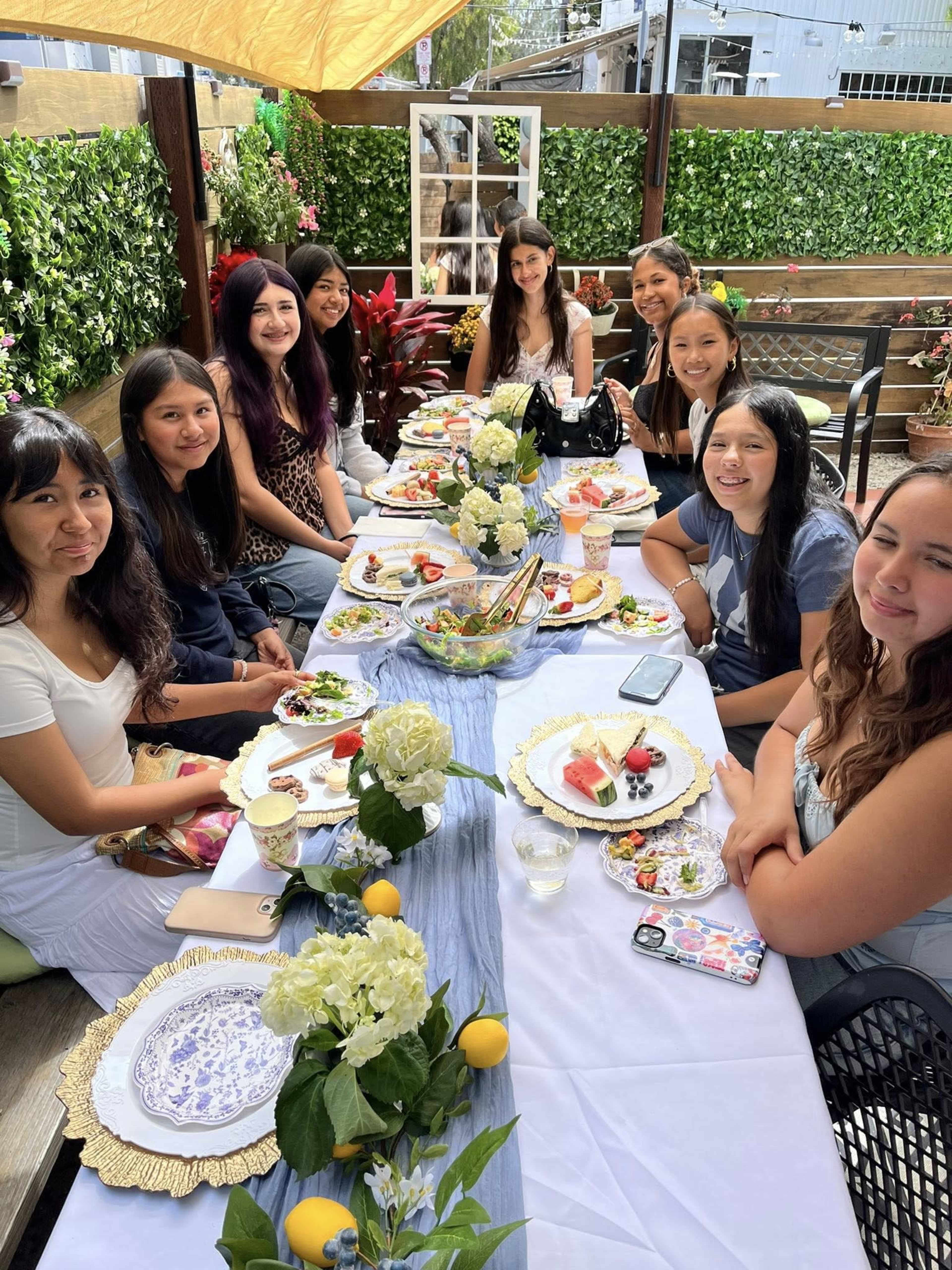 A group of eleven people sits around a long table set with food and drinks in an outdoor garden setting.