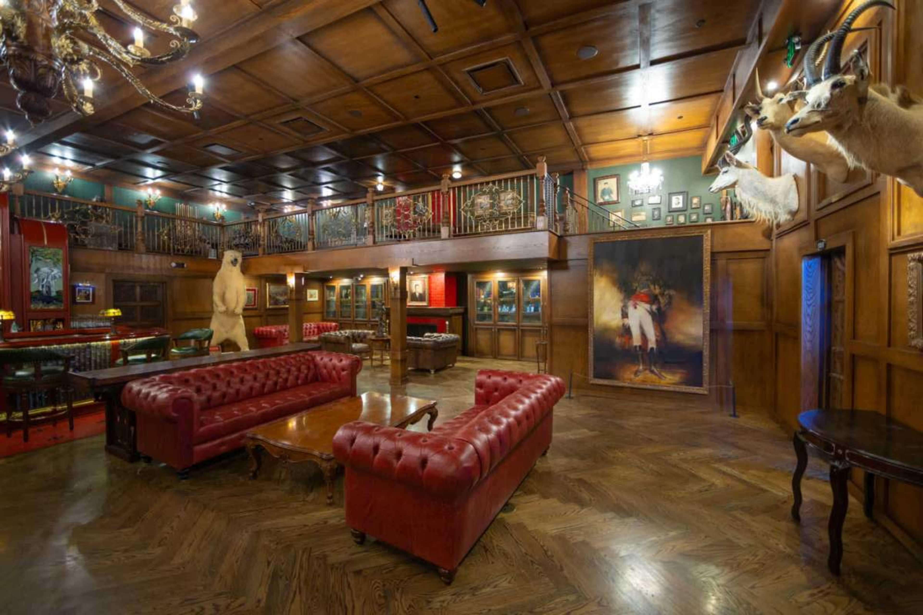 The image shows a rustic room featuring dark wood paneling, a large bear statue, mounted animal heads, and red leather seating.