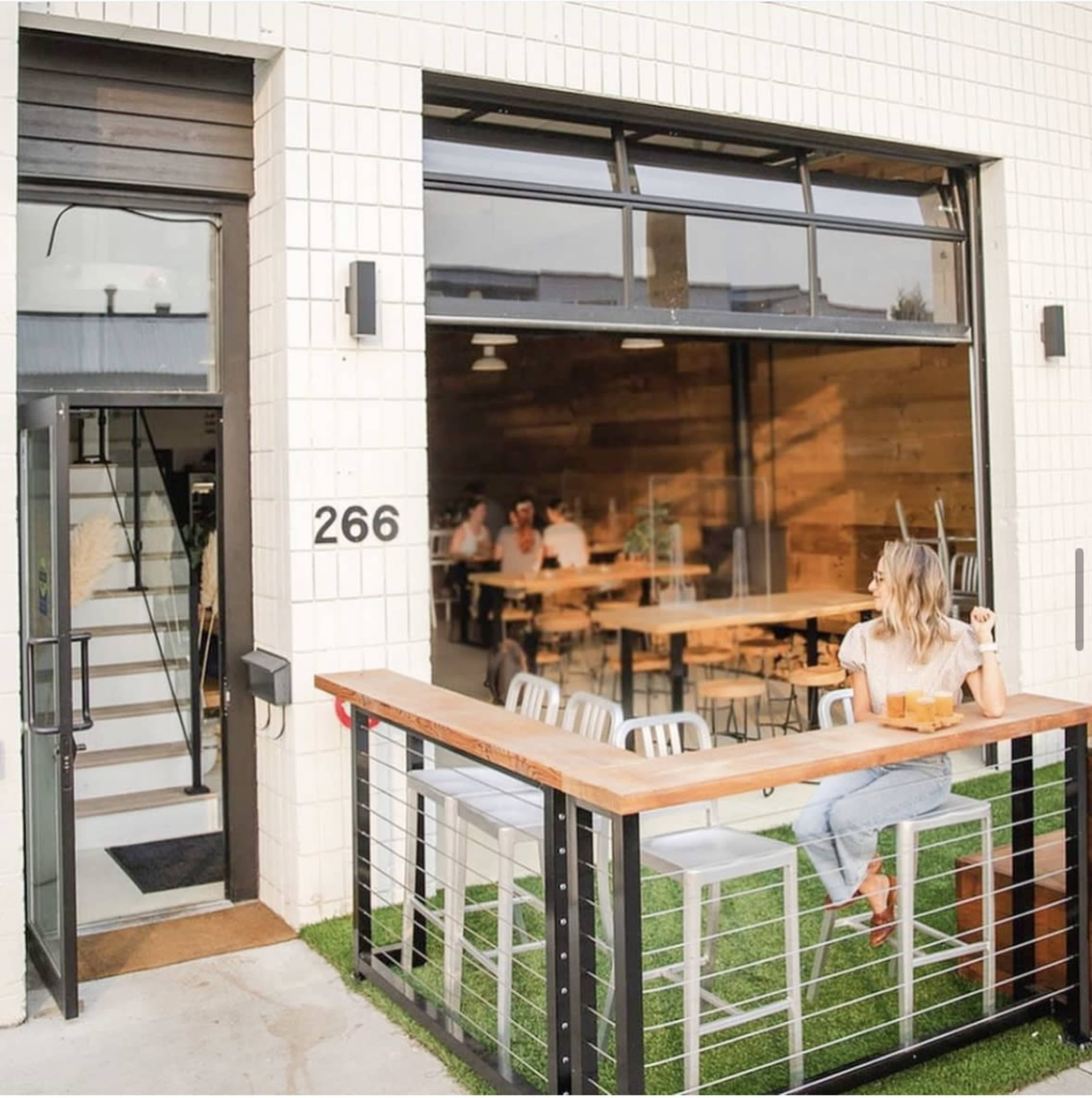 A woman sits at a wooden bar table outside a restaurant with an open front, while patrons are visible inside.