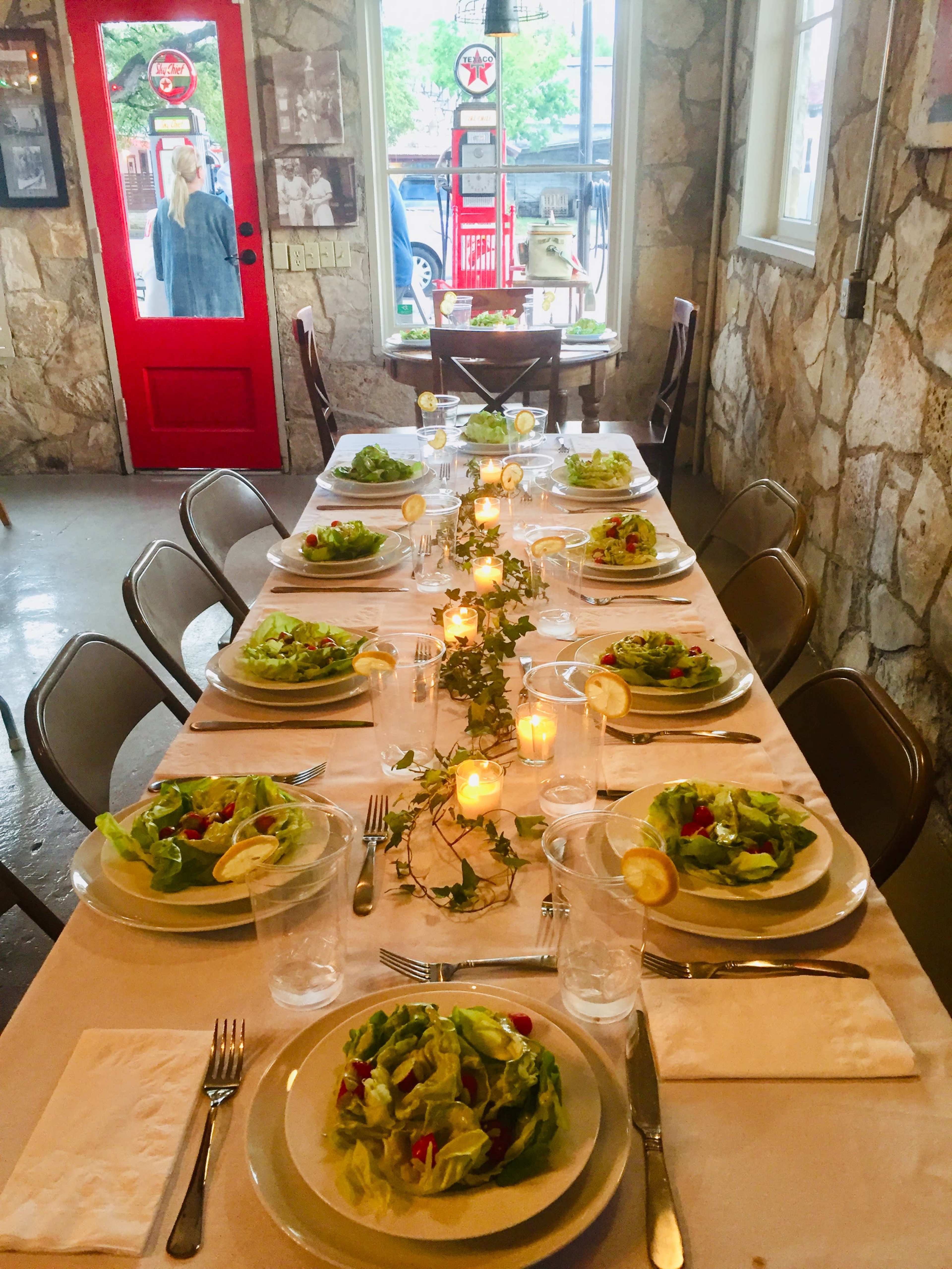 A long dining table is set with salads, plates, glasses, and candles, lined with greenery, against a stone wall and a red door.