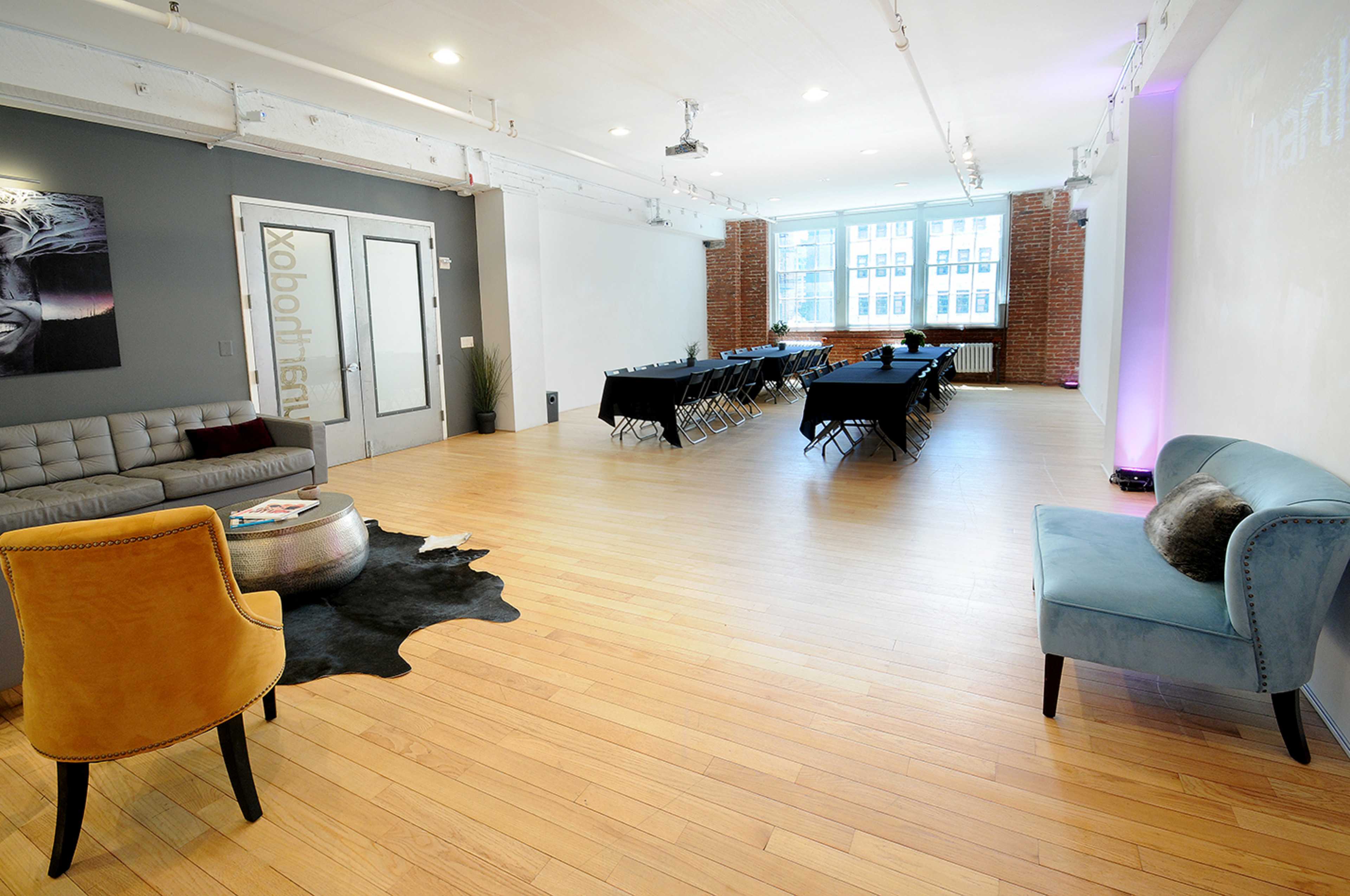 A modern event space features a seating area with a gray couch and yellow chair, along with a central arrangement of tables and chairs ready for a meeting.