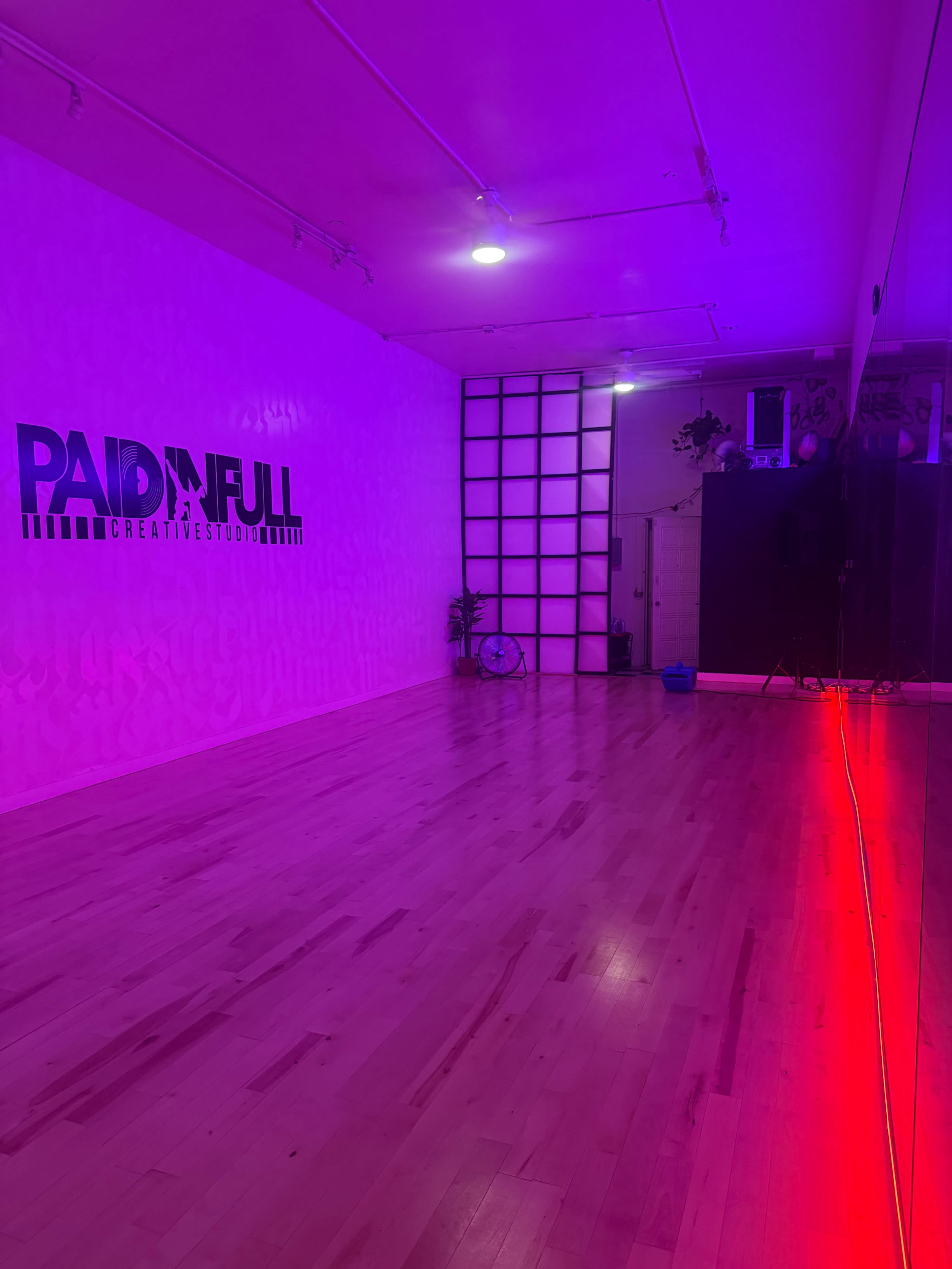 The image shows a spacious, brightly lit studio featuring a mirrored wall, hardwood flooring, and a vibrant pink and purple lighting scheme.