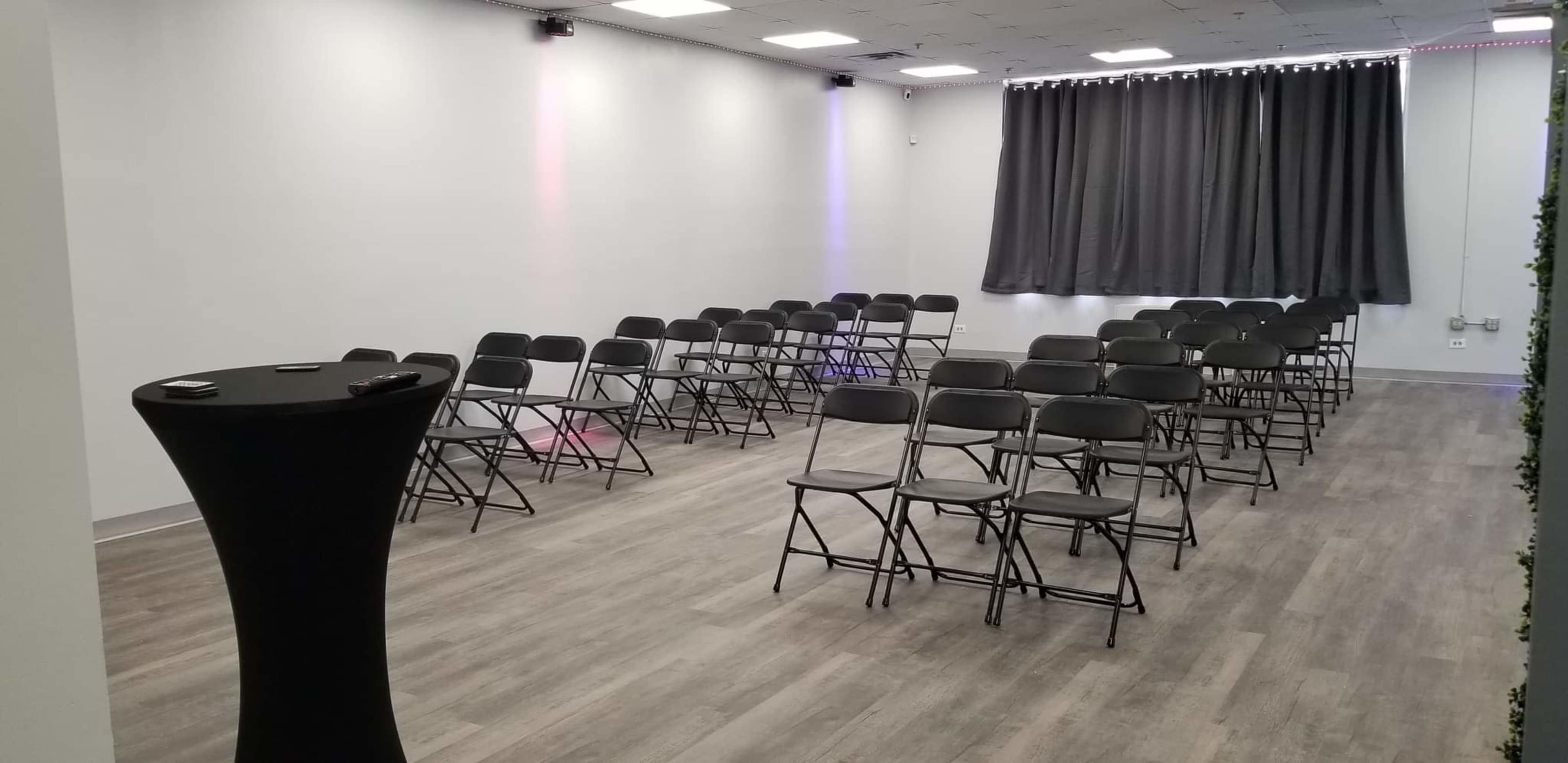 A sparsely furnished room with rows of black folding chairs facing a central point and a small table off to the side.