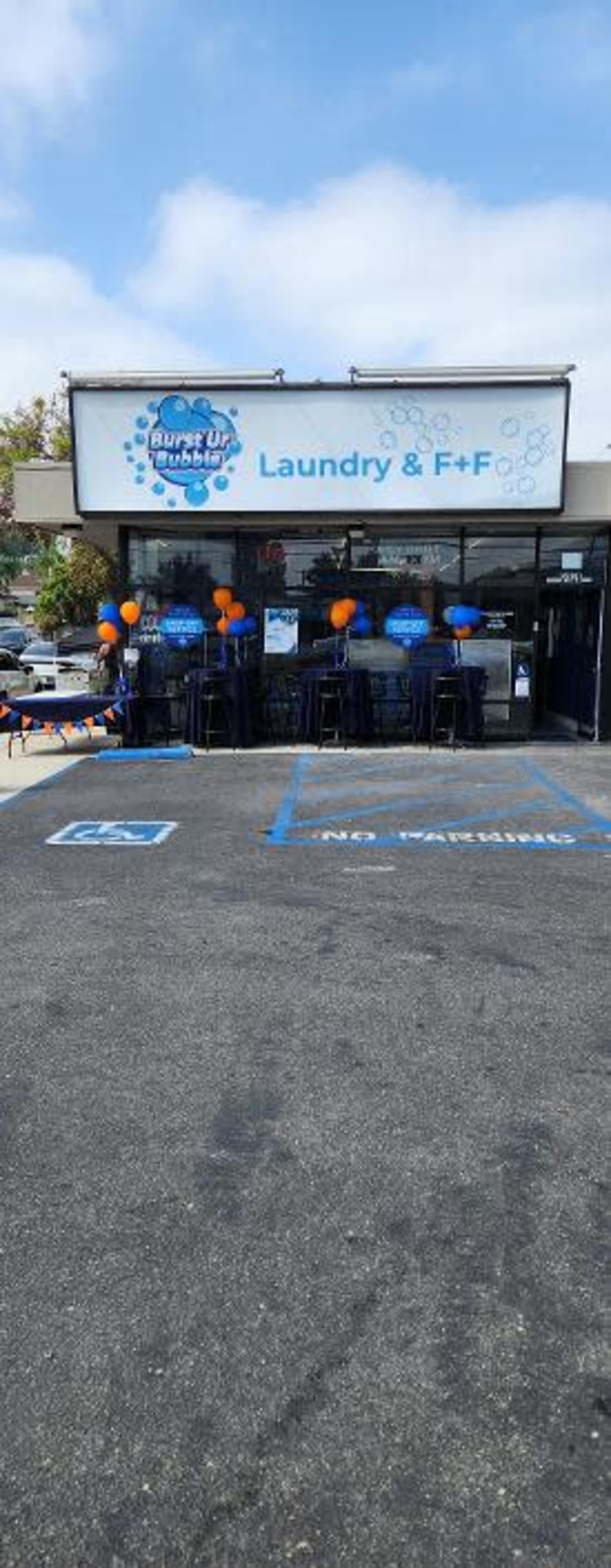 The image shows the exterior of a laundry business called "Super Clean Laundry & F+F," featuring a colorful sign and decorated with balloons in orange and blue.