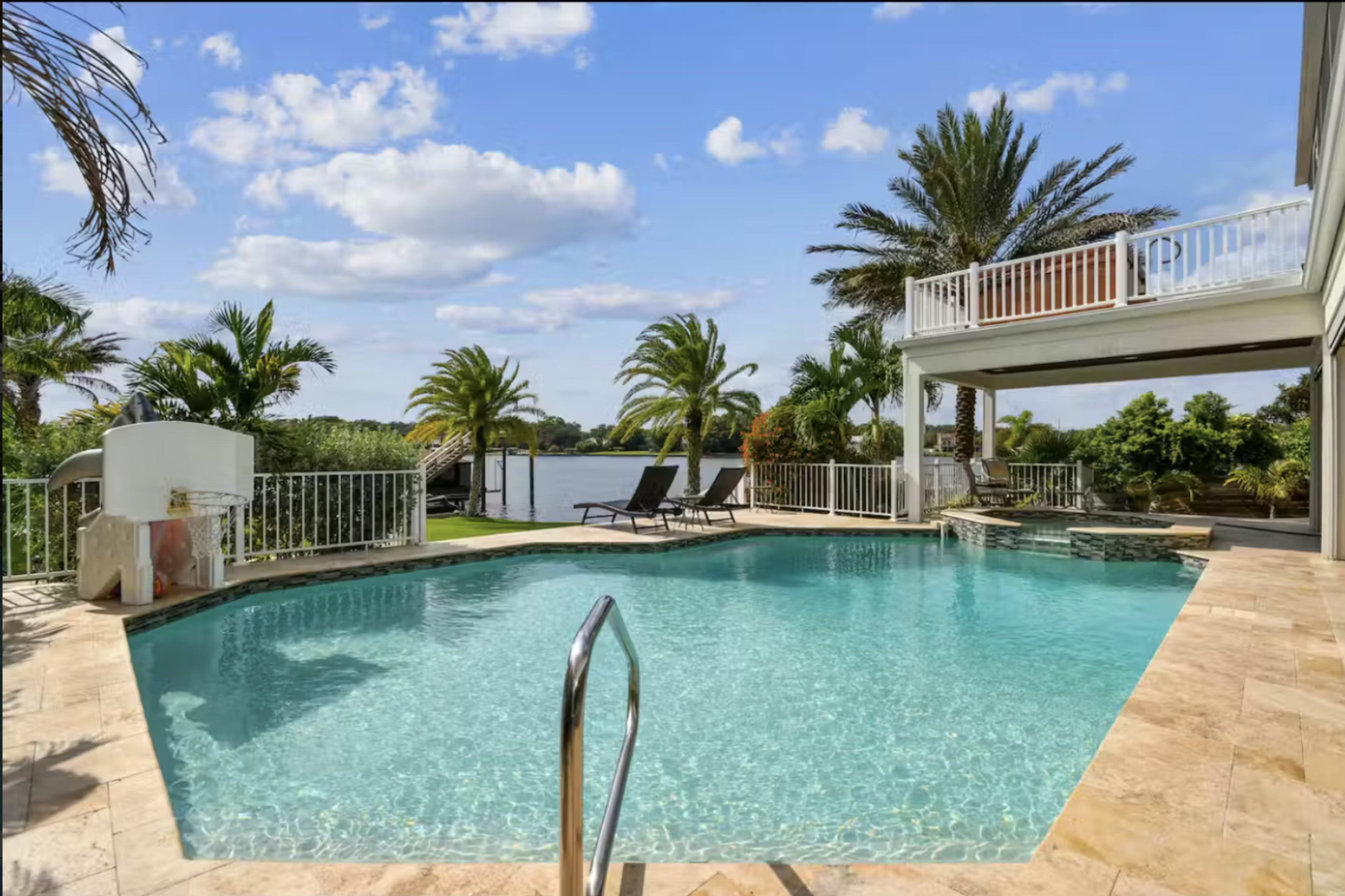A swimming pool with a clear blue surface is surrounded by palm trees and a patio, overlooking a waterway.