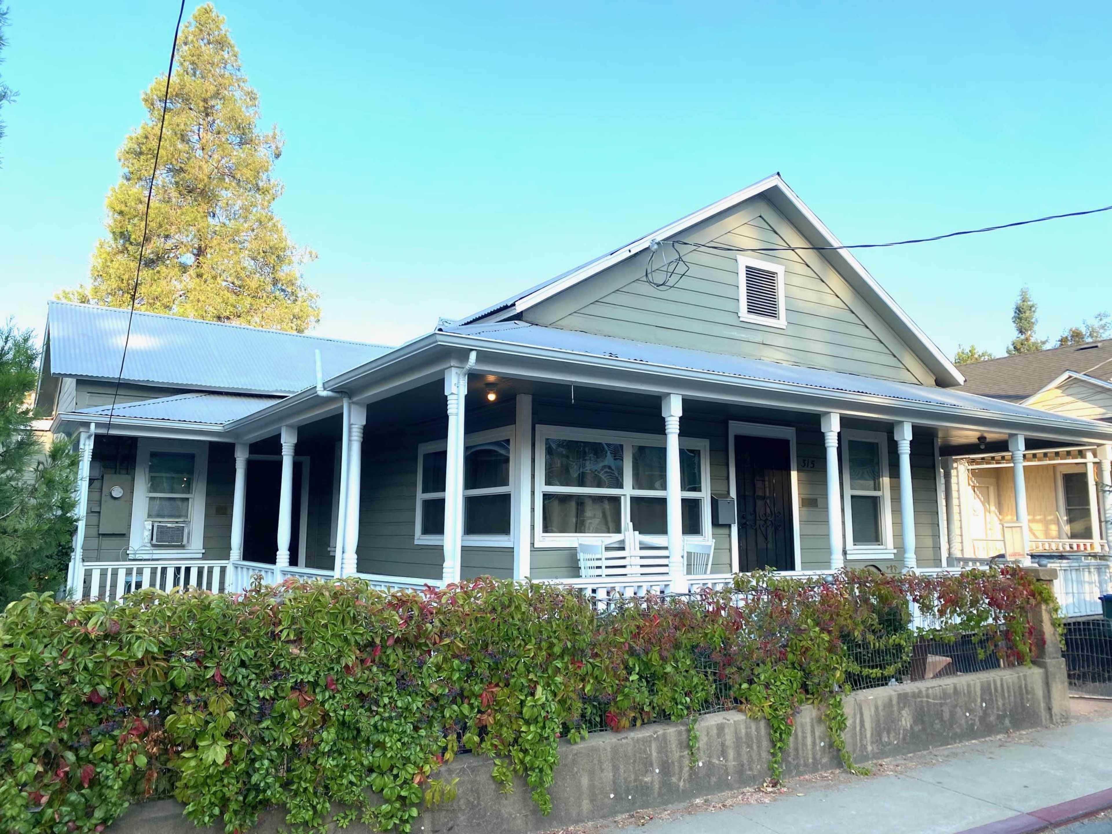 A light green house with a porch surrounded by shrubs and a pathway, set against a clear blue sky.
