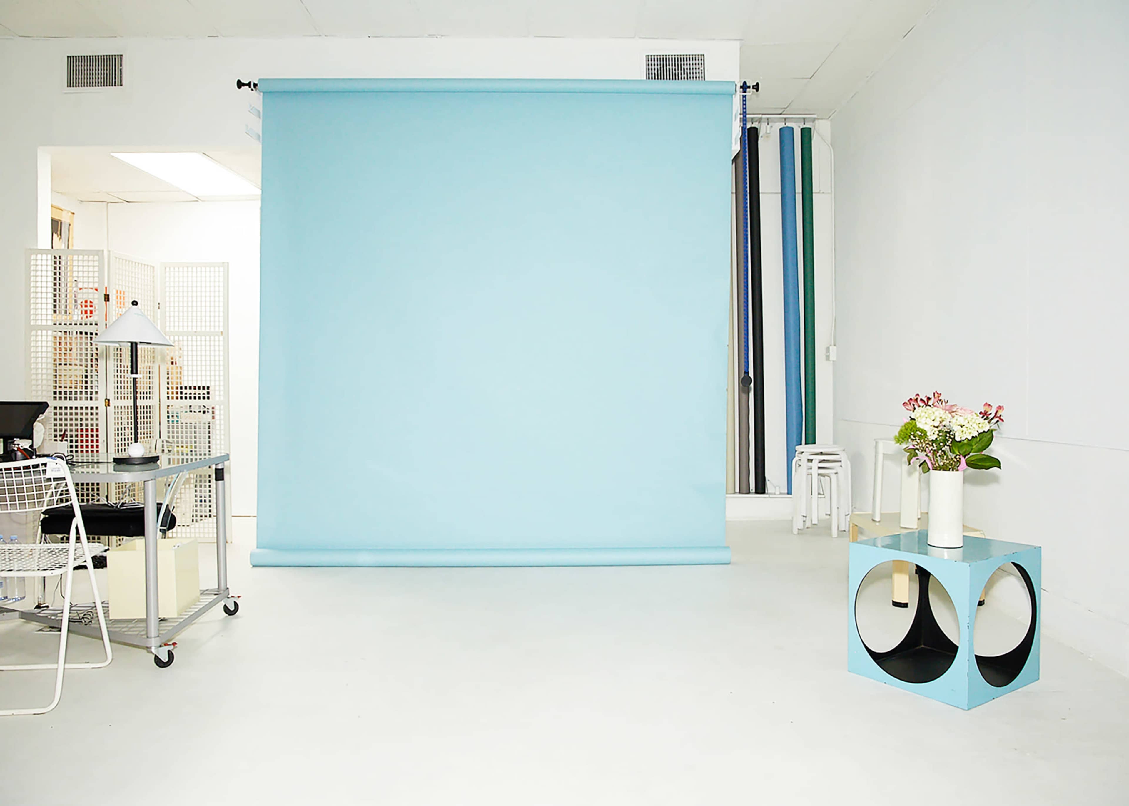 A bright white room with a pale blue backdrop, a small blue table, and a workstation featuring a lamp and various studio equipment.