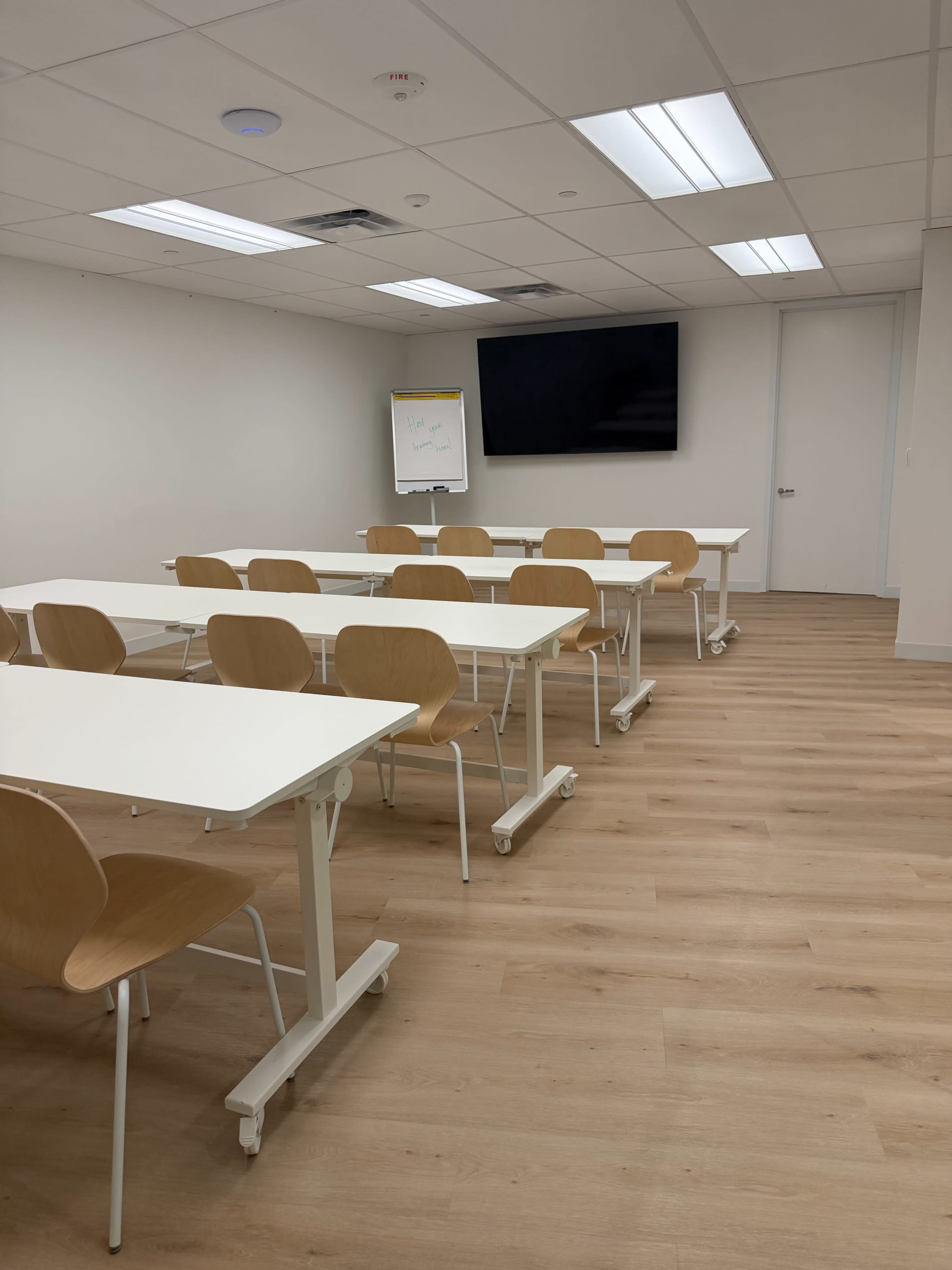 The image shows a modern classroom with several white tables and wooden chairs arranged in rows, a flip chart, and a large screen on the wall.