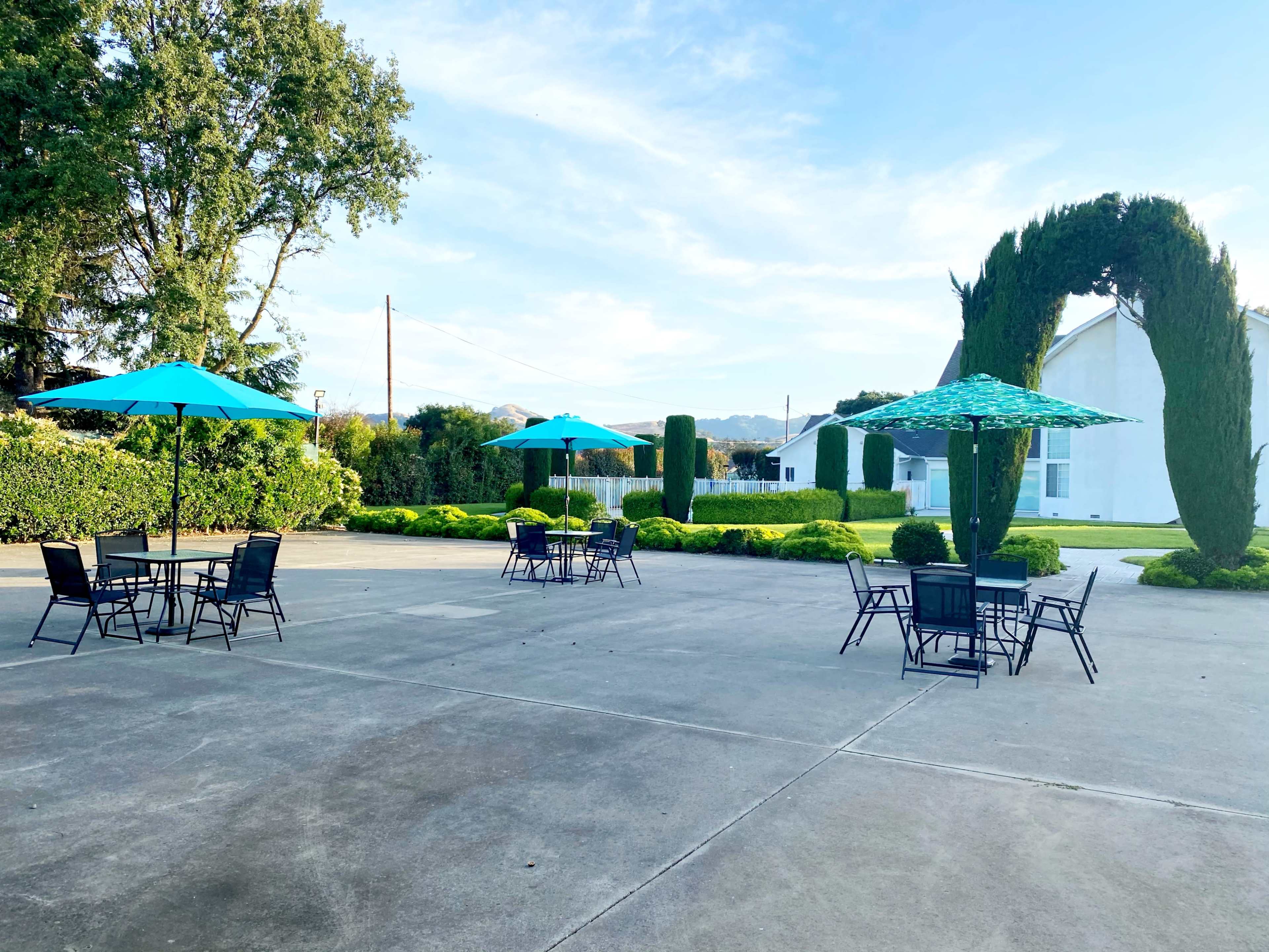 The image shows a paved outdoor area with several tables and turquoise umbrellas, surrounded by neatly trimmed hedges and trees.