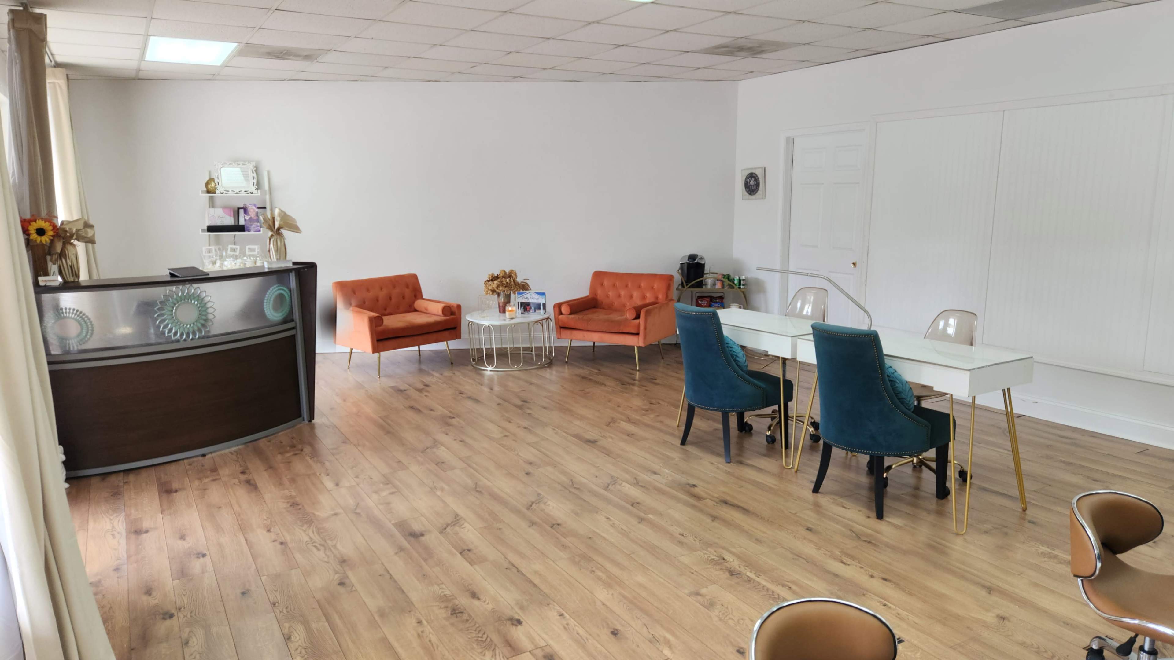 The image shows a spacious room with a reception area featuring a wooden desk, two orange chairs, a glass table, and several turquoise chairs around a white dining table.