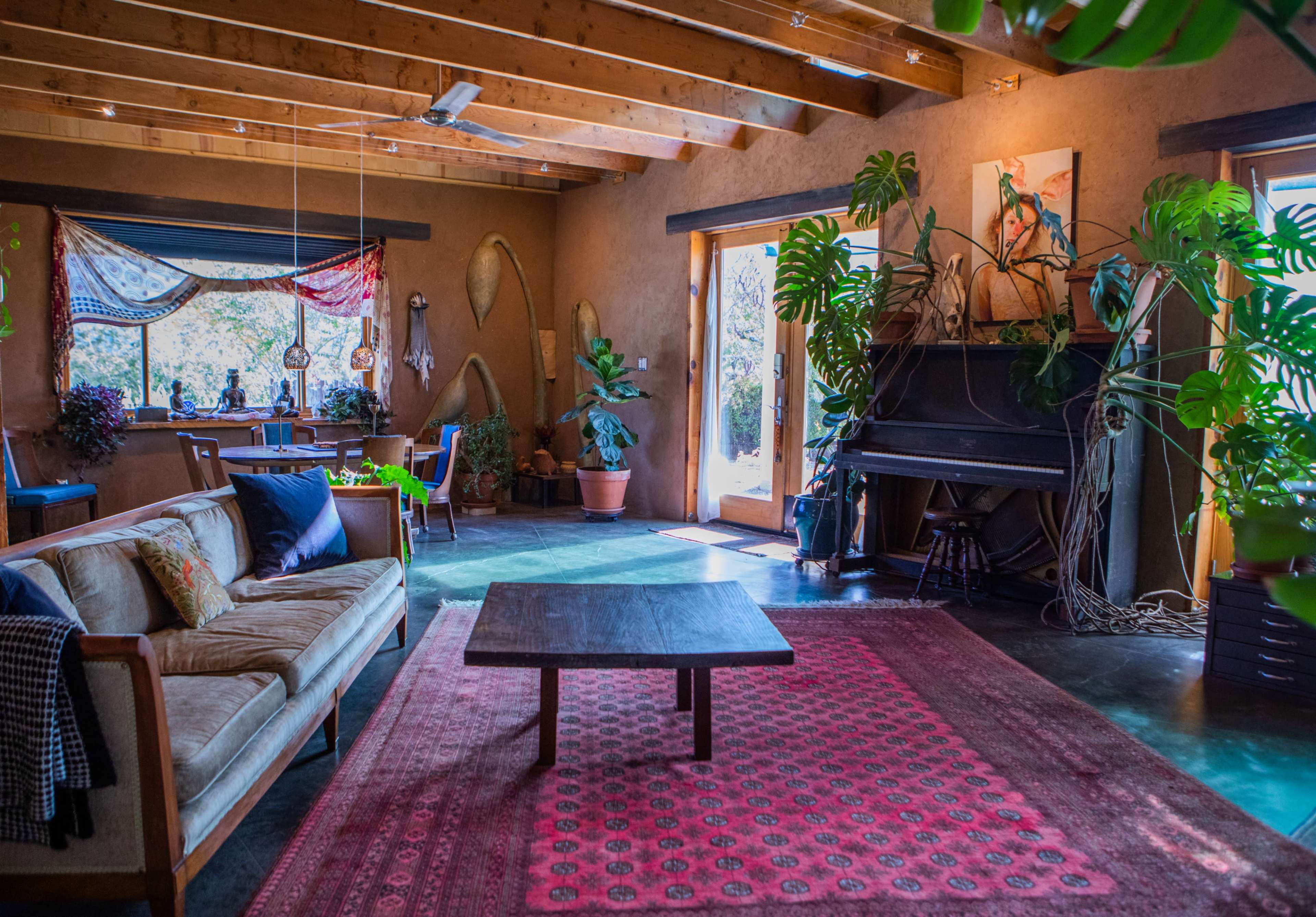 The image shows a cozy living room with a wooden ceiling, a sofa, a coffee table, plants, and a piano near large windows.