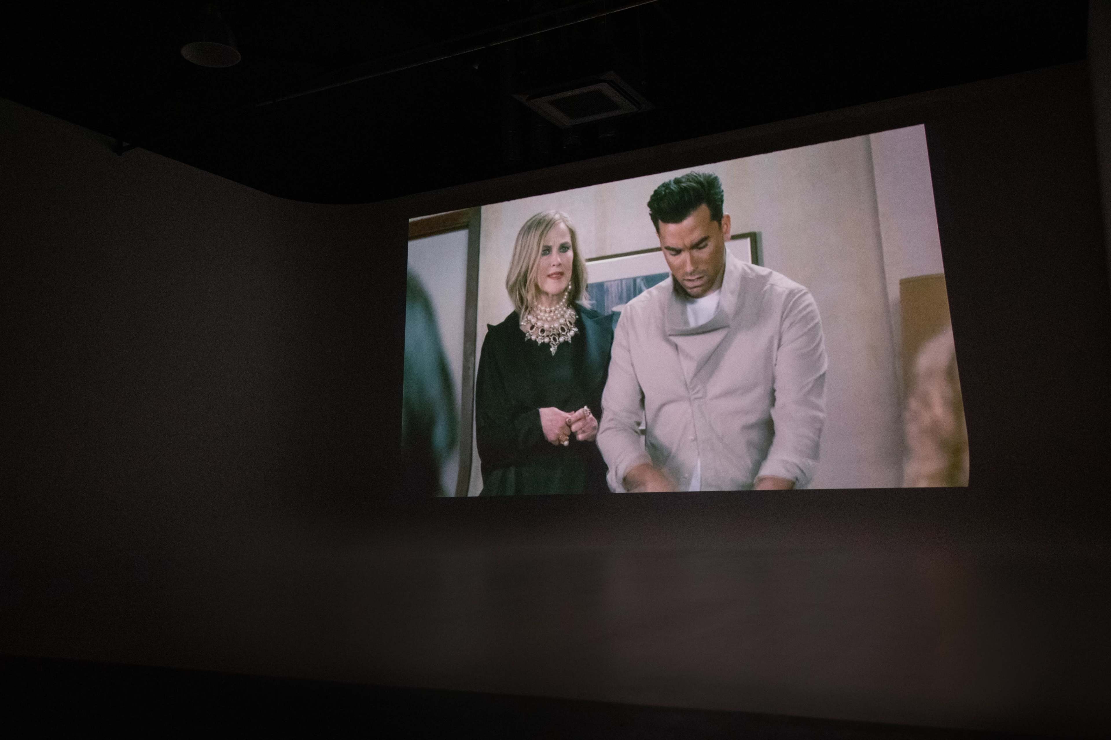 A large projection shows a scene featuring two individuals, a man wearing a chef's uniform and a woman in a dark outfit, in an indoor setting.