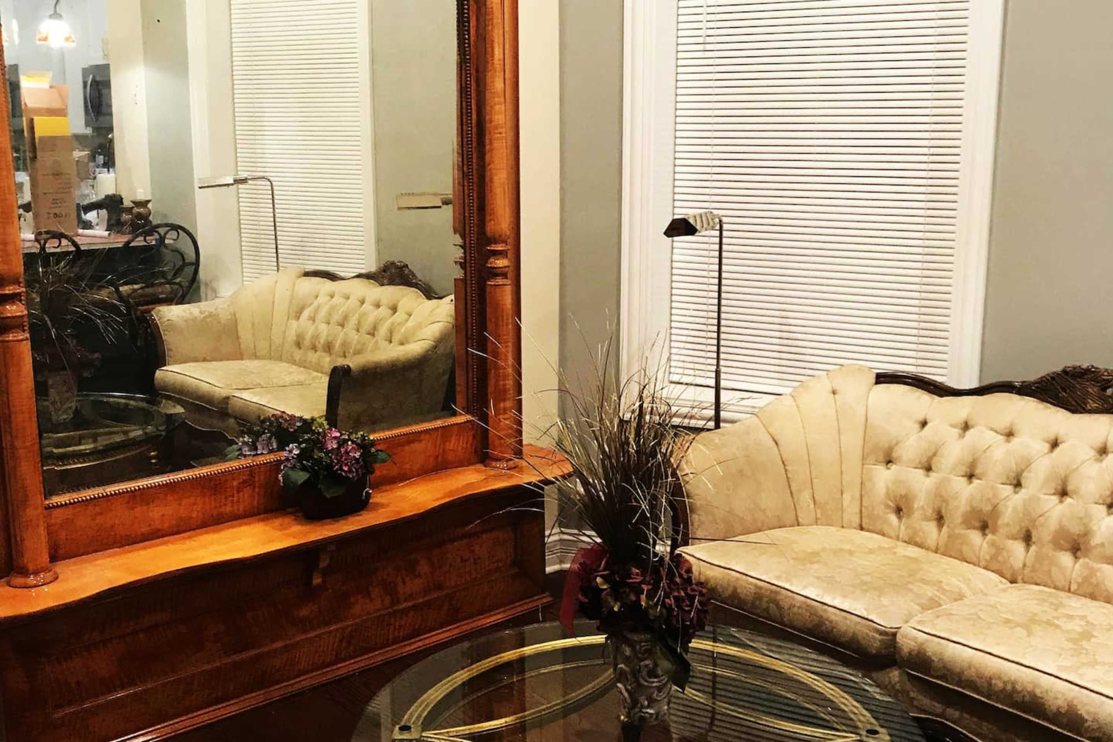 The image shows a cozy sitting area featuring a tufted beige sofa, a round glass coffee table, and a large wooden mirror reflecting the space.
