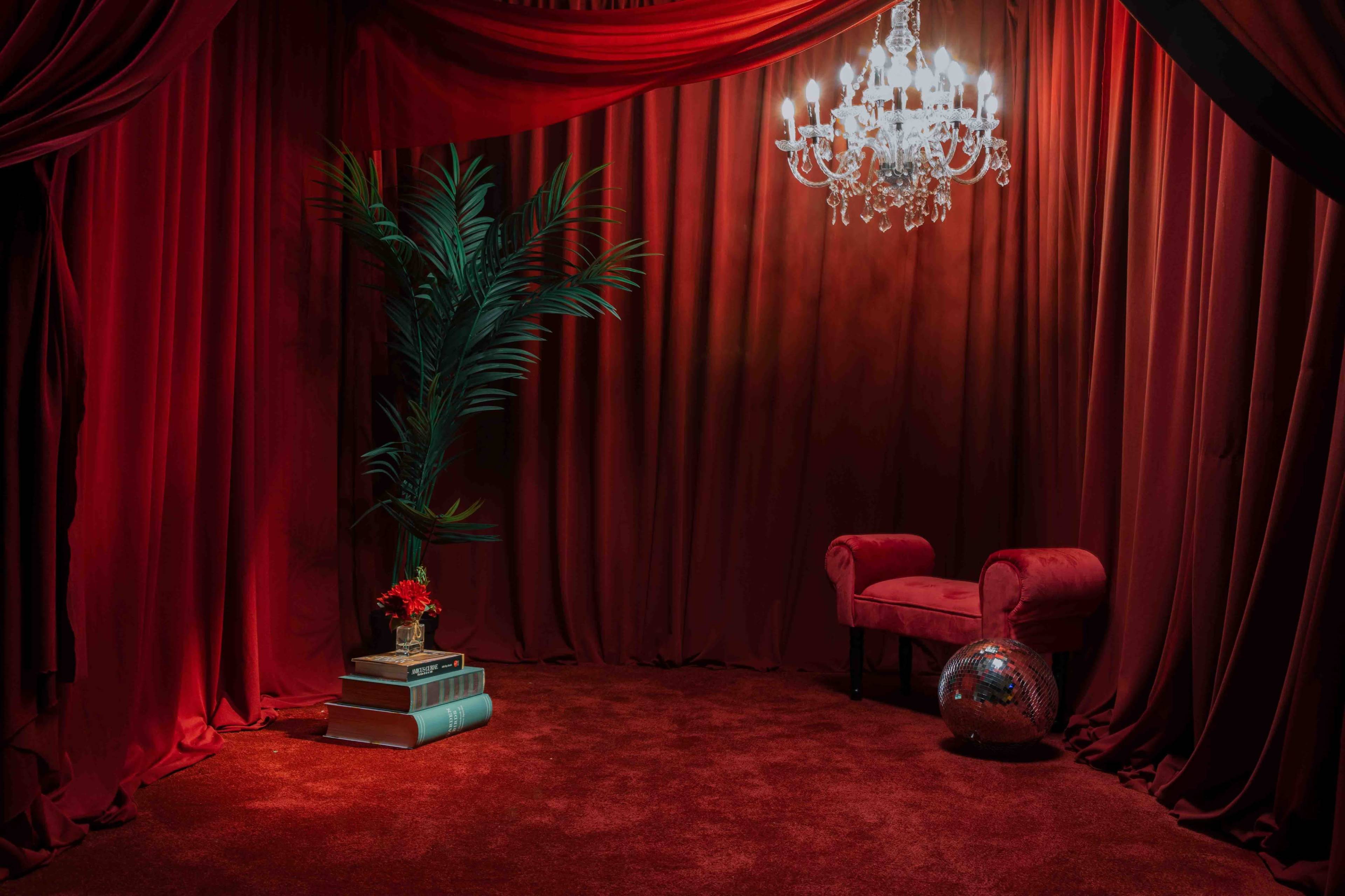 A red velvet curtain backdrop features a plush armchair, a stack of books, a potted plant, and a chandelier overhead.