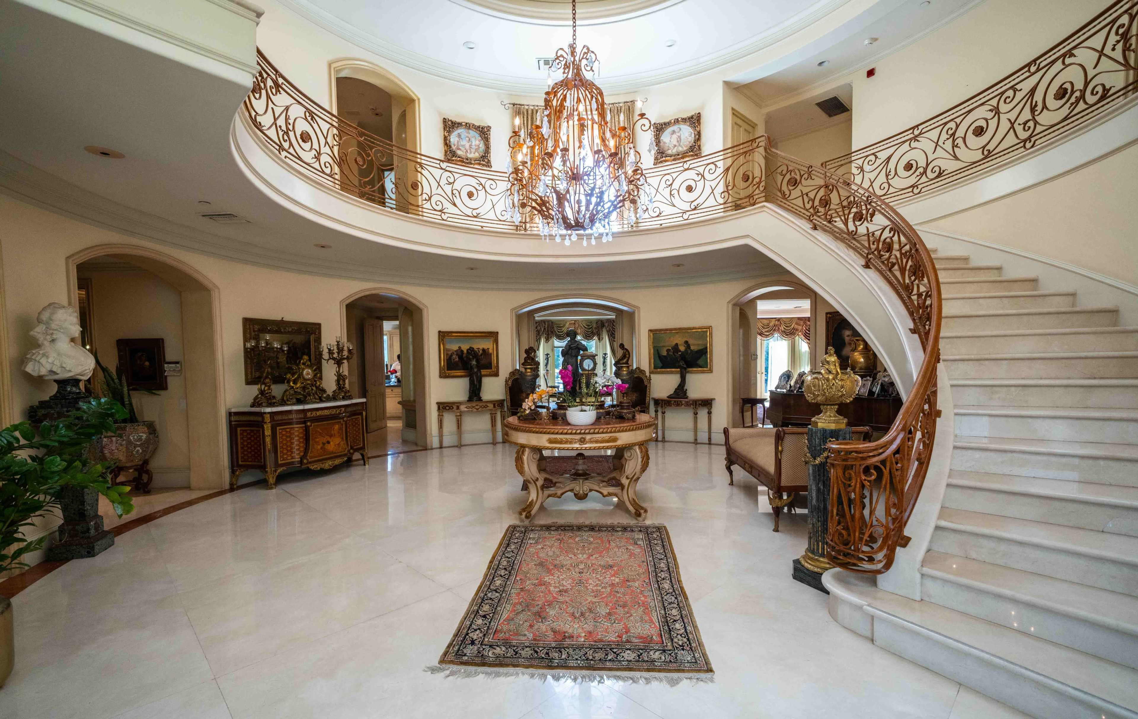 The image shows an elegant two-story foyer with a grand staircase, a chandelier, and ornate furnishings.