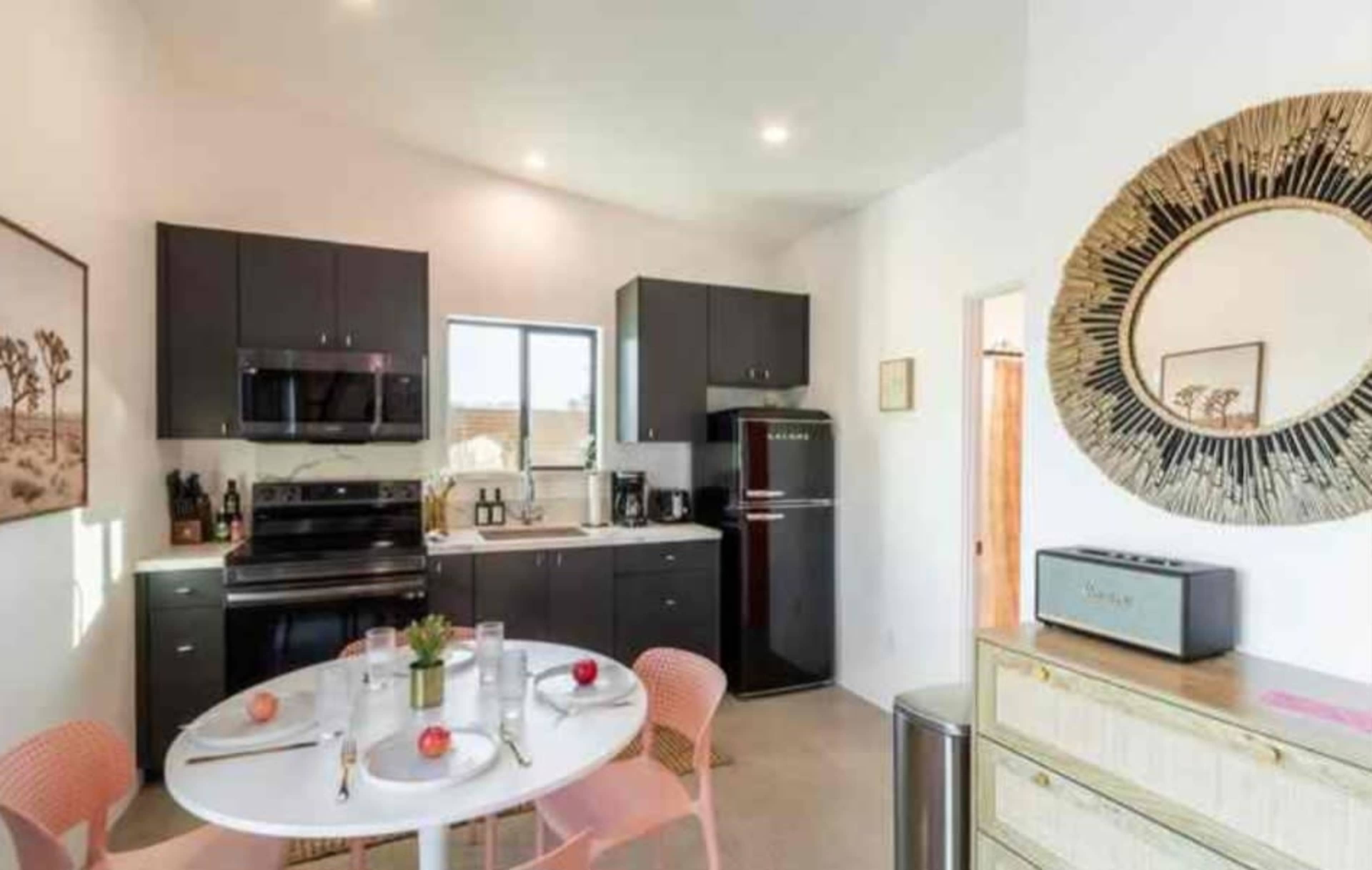 The image shows a modern kitchen featuring black cabinets, stainless steel appliances, a round dining table with pink chairs, and a circular wall mirror.