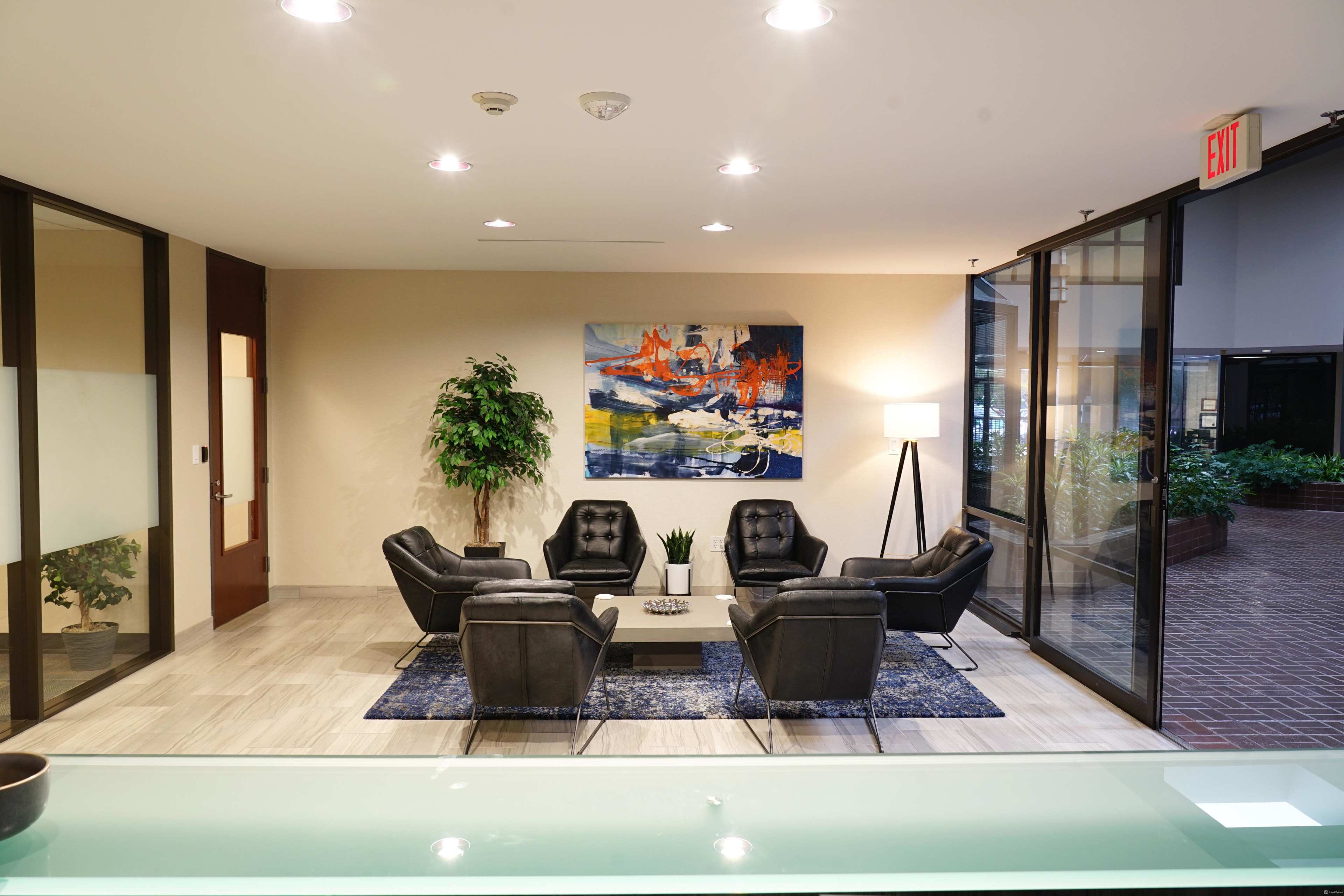 A modern lounge area featuring four black armchairs arranged around a coffee table, with a large abstract painting on the wall and a potted plant in the corner.