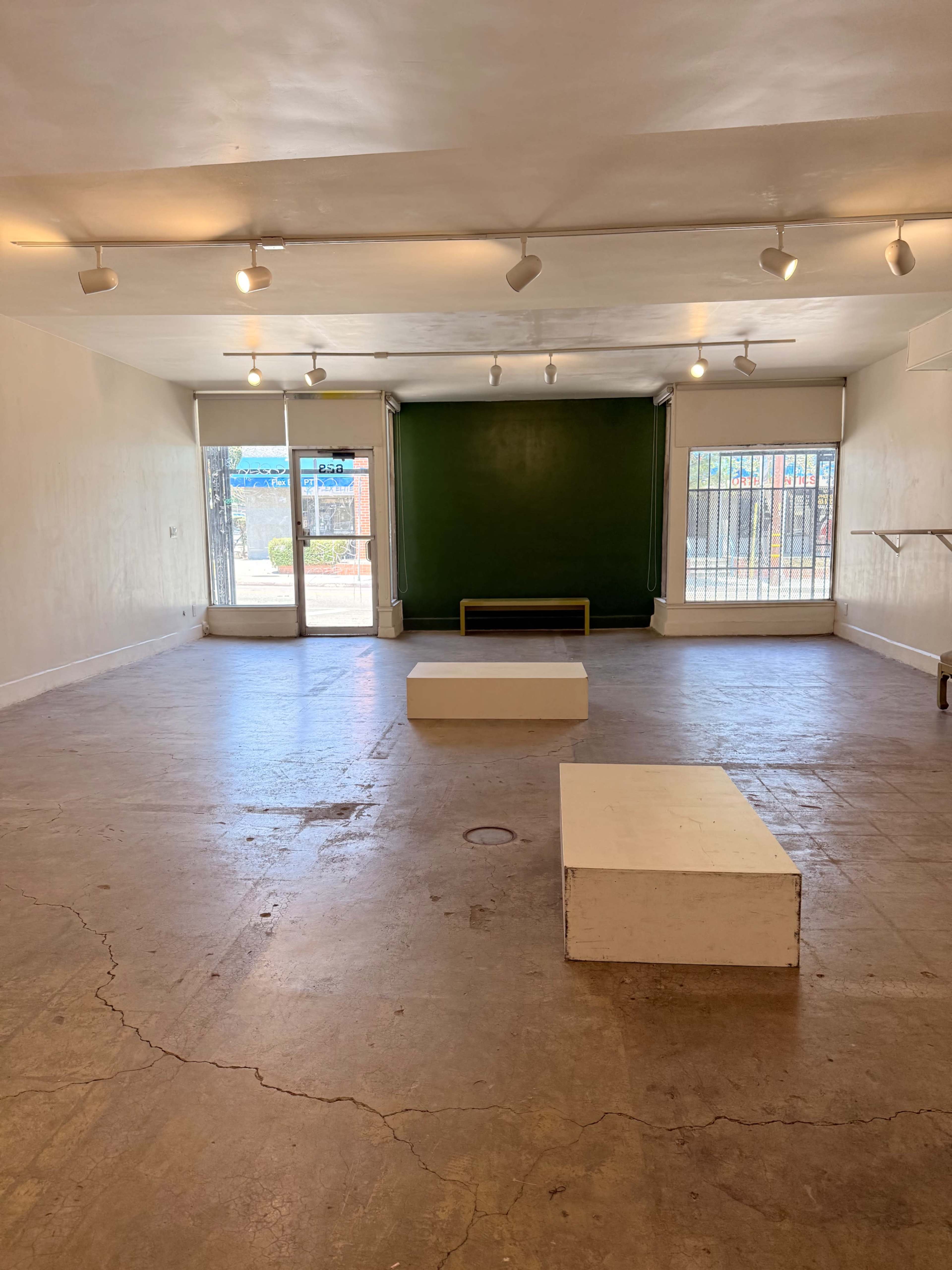 The image depicts an empty art gallery space with two white display platforms and a green backdrop at one end.