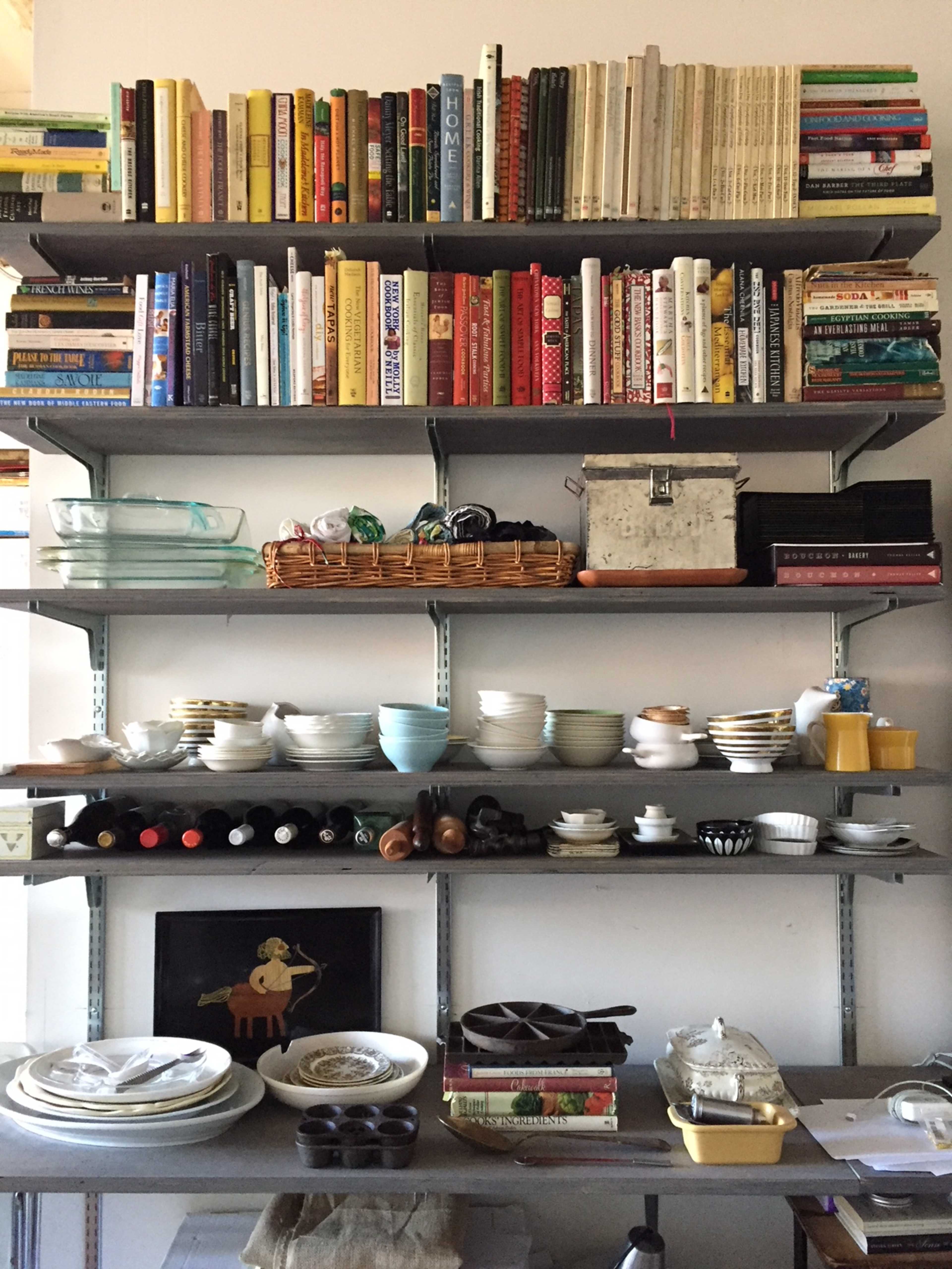 A bookshelf filled with various books above a countertop displaying an assortment of dishes, cookware, and storage containers.