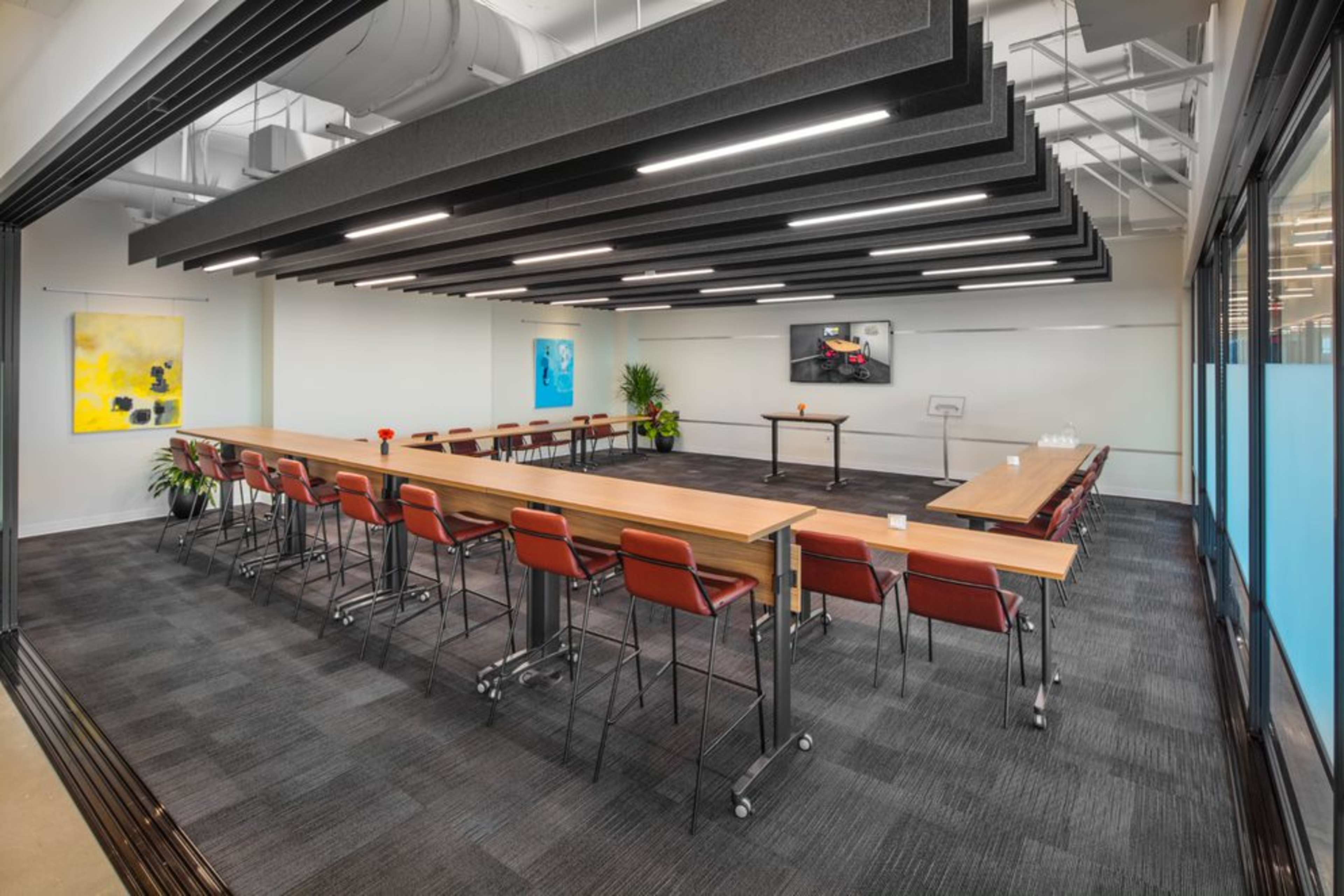 A modern conference room with a long rectangular table, red chairs, and a presentation screen at the front.