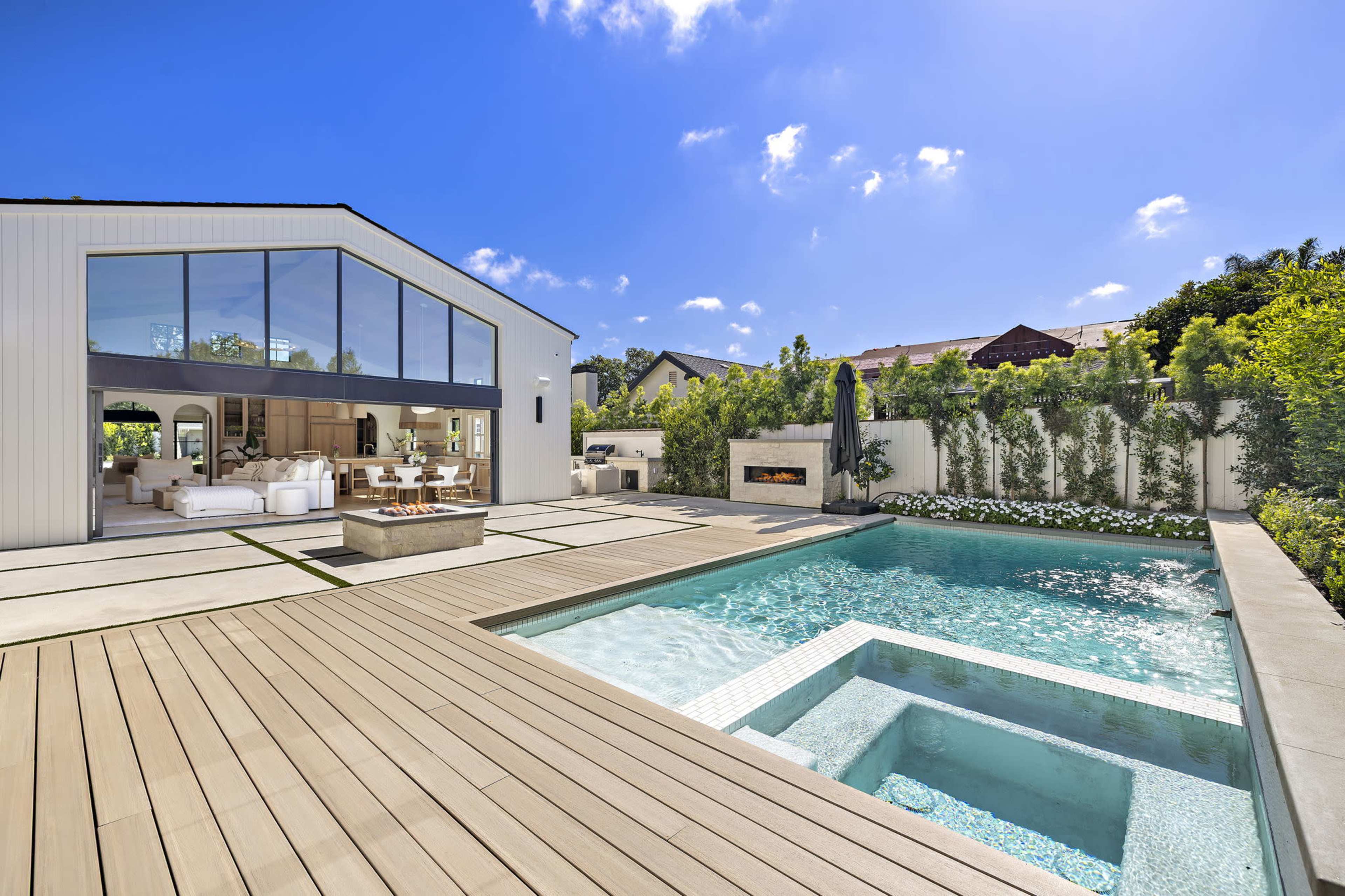 The image shows a modern home with large windows, a swimming pool featuring a spa area, and a wooden deck surrounded by greenery.