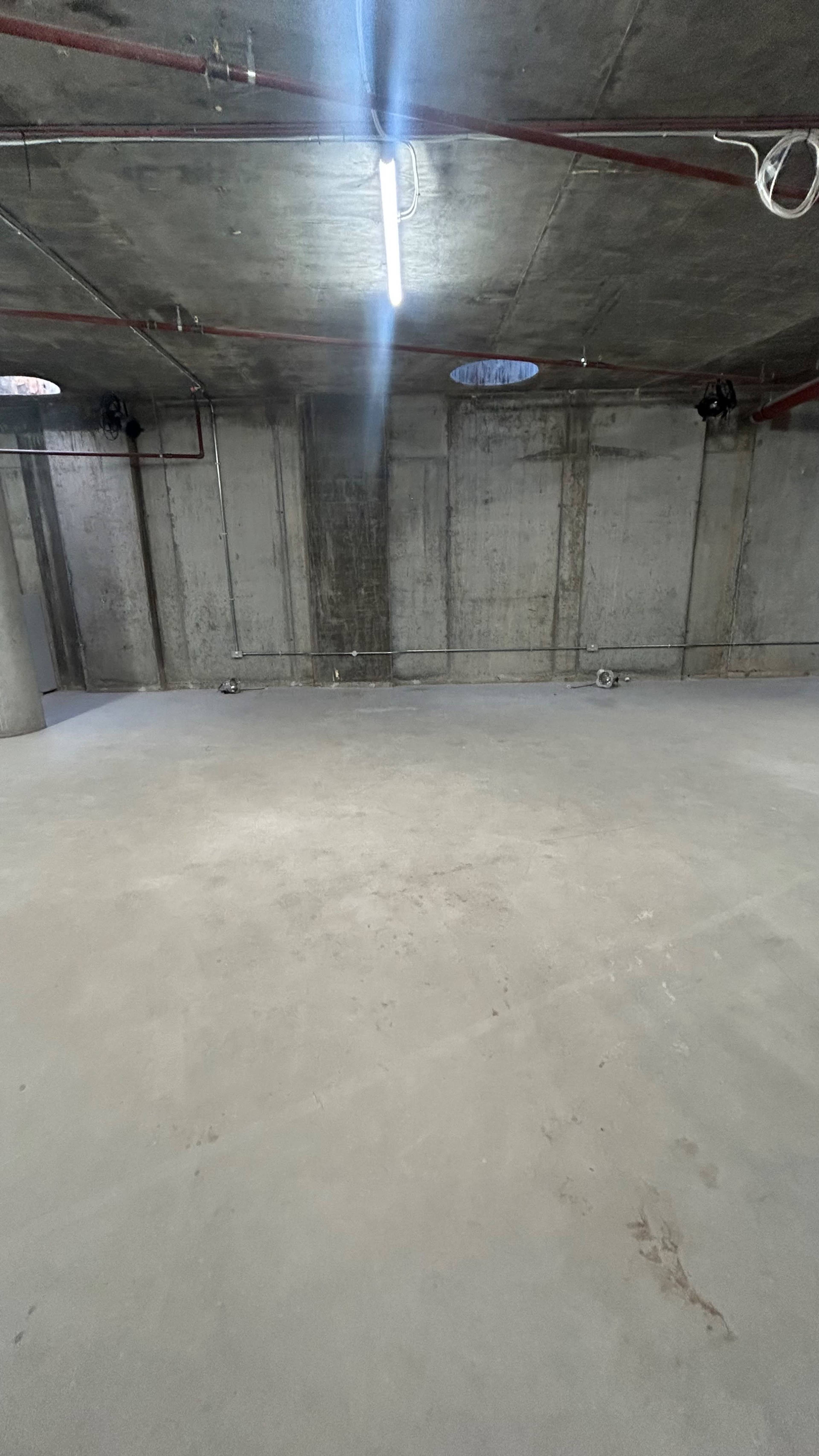 The image shows a large, empty concrete room with exposed walls and a ceiling, illuminated by overhead fluorescent lights.