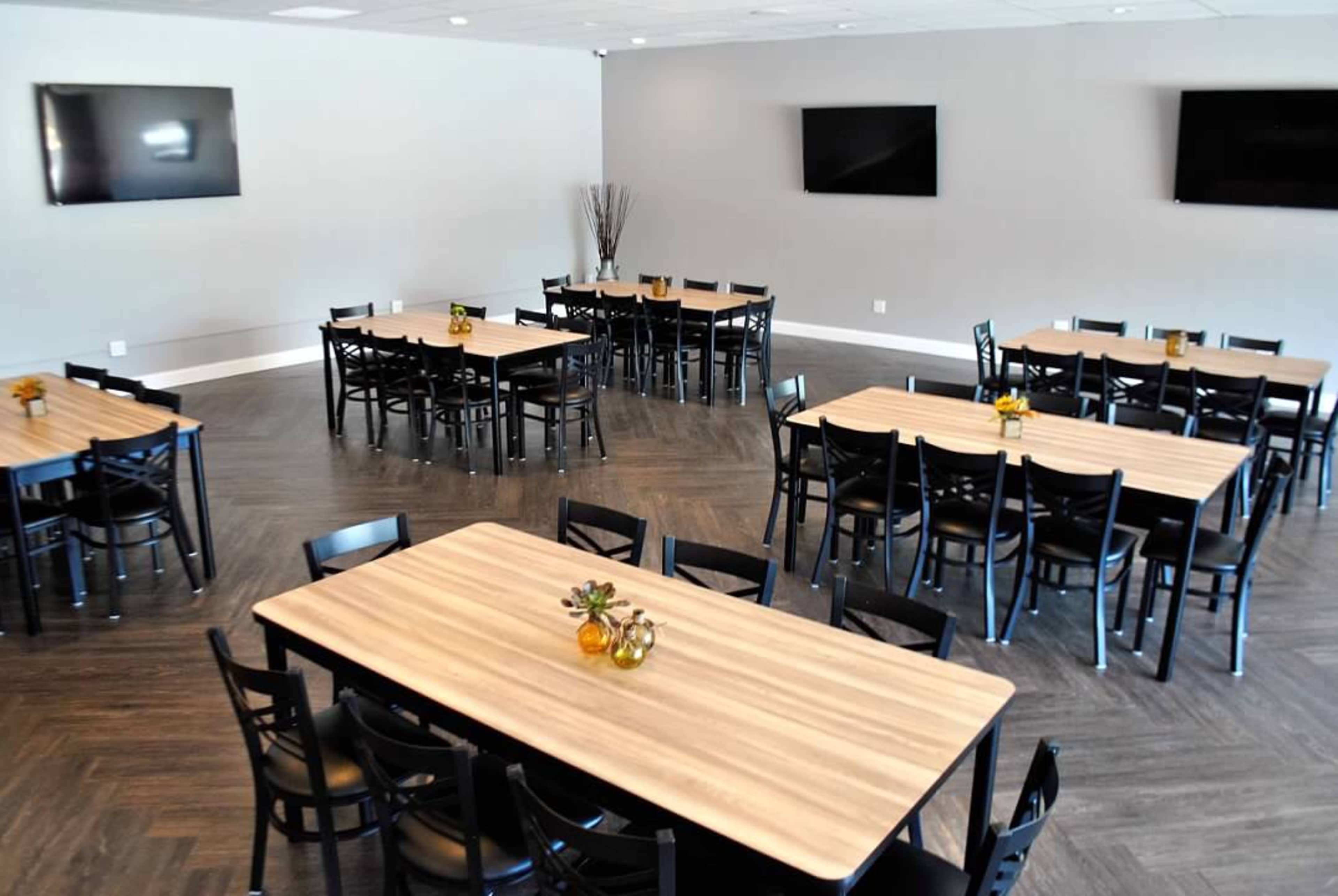 A spacious room arranged with multiple tables and chairs, featuring a modern design and two flat-screen televisions on the walls.