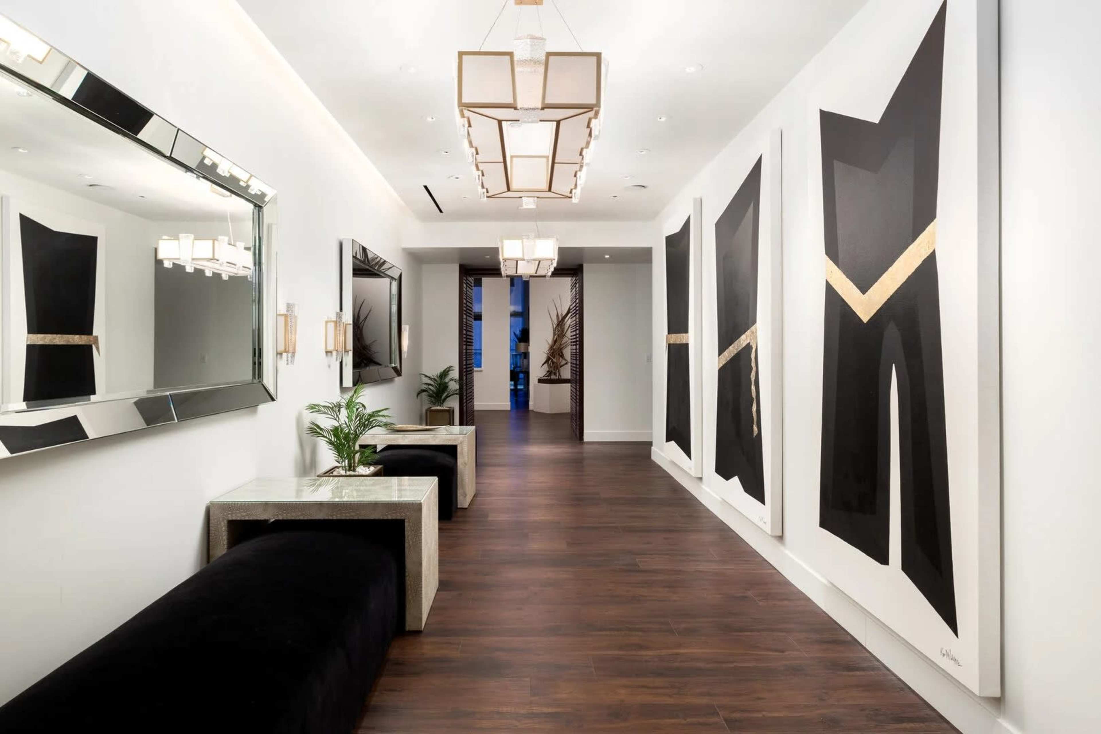 The image shows a modern hallway featuring large black and white abstract paintings, a mirror, elegant lighting fixtures, and minimalist seating areas.