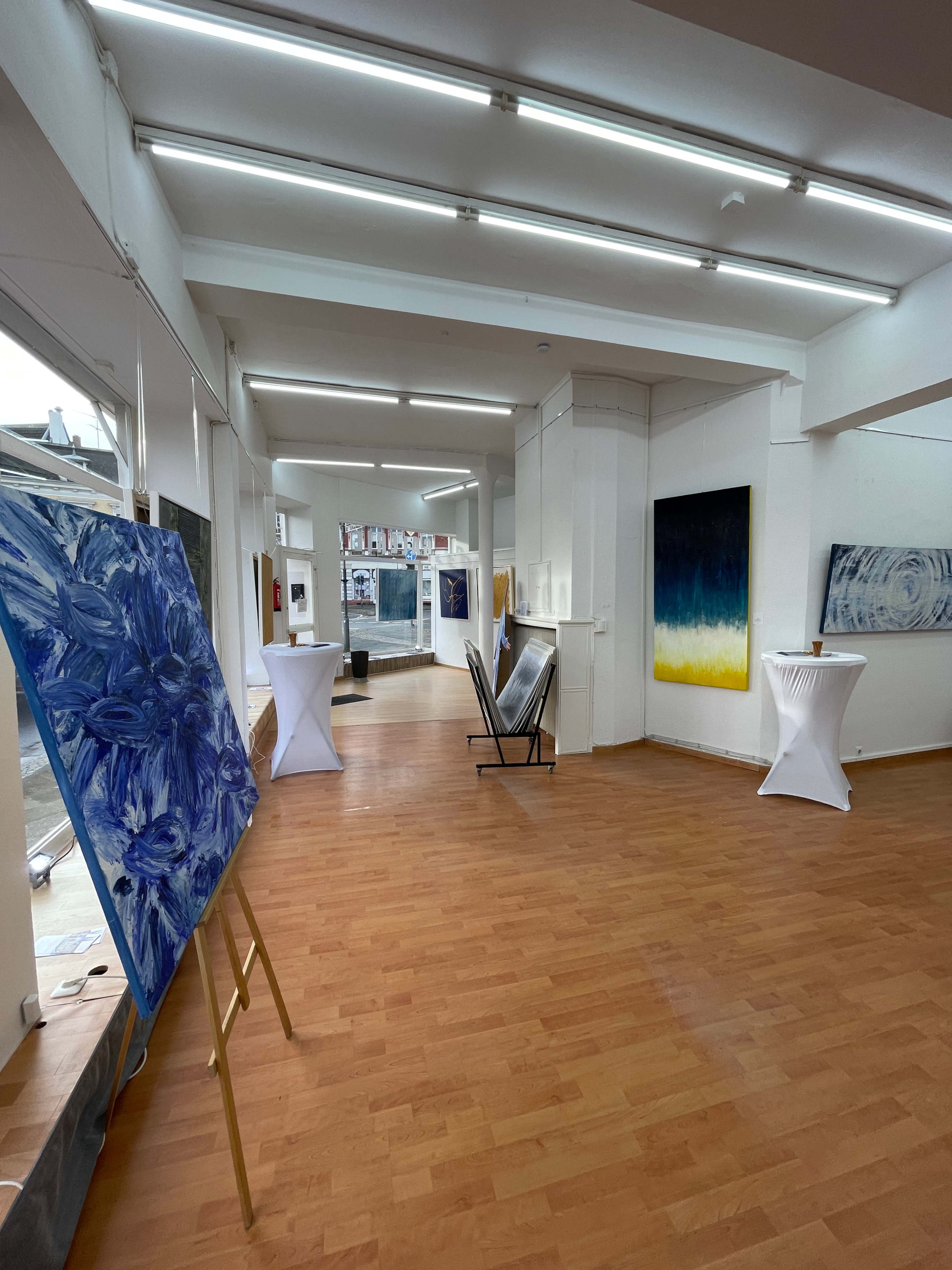 A spacious art gallery featuring several large paintings displayed on easels and wall-mounted, with white tablecloth-covered tables set up in the center.