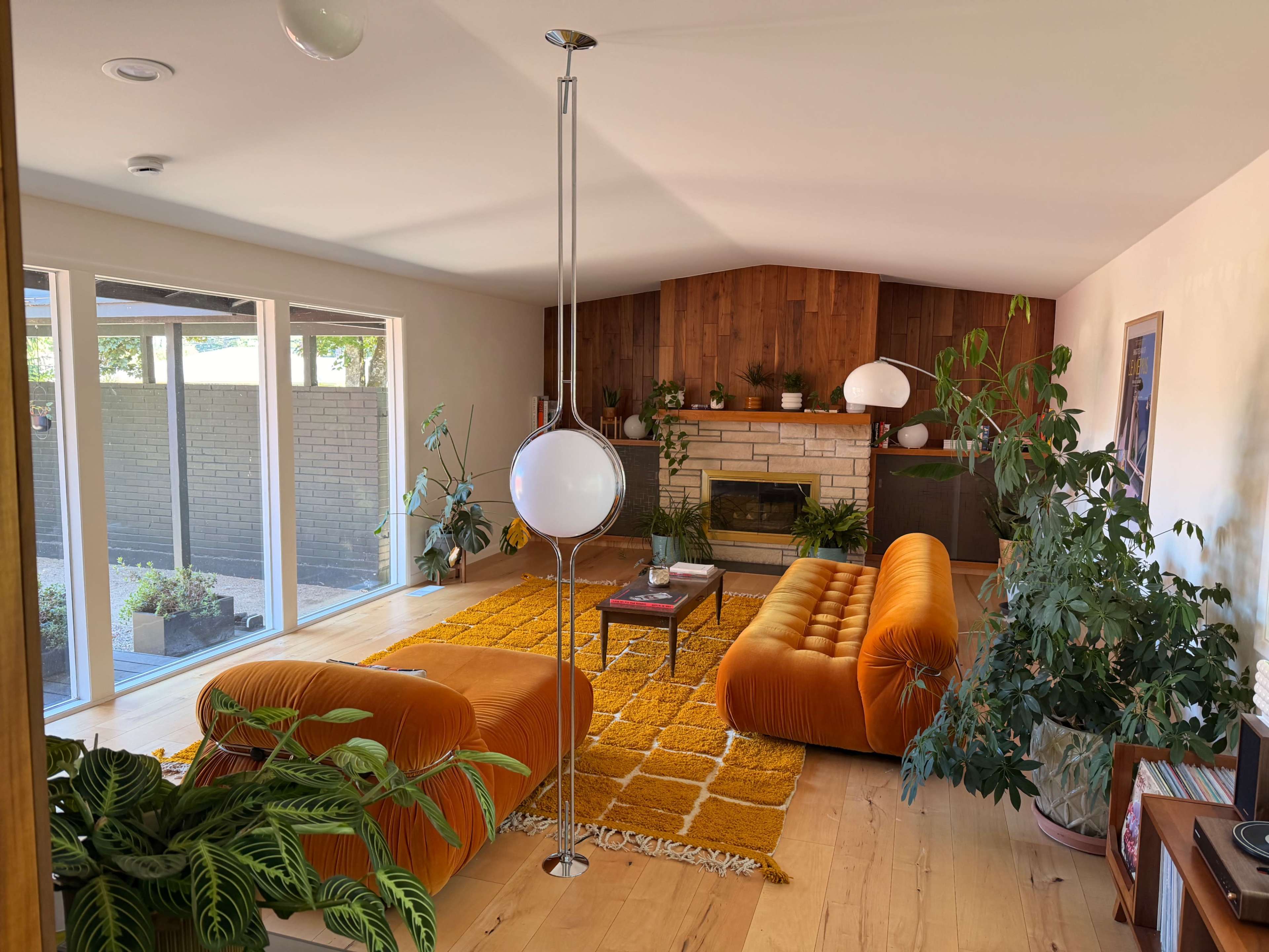 The scene features a cozy living room with two orange sofas, a wooden accent wall, a fireplace, large windows allowing natural light, and various indoor plants.