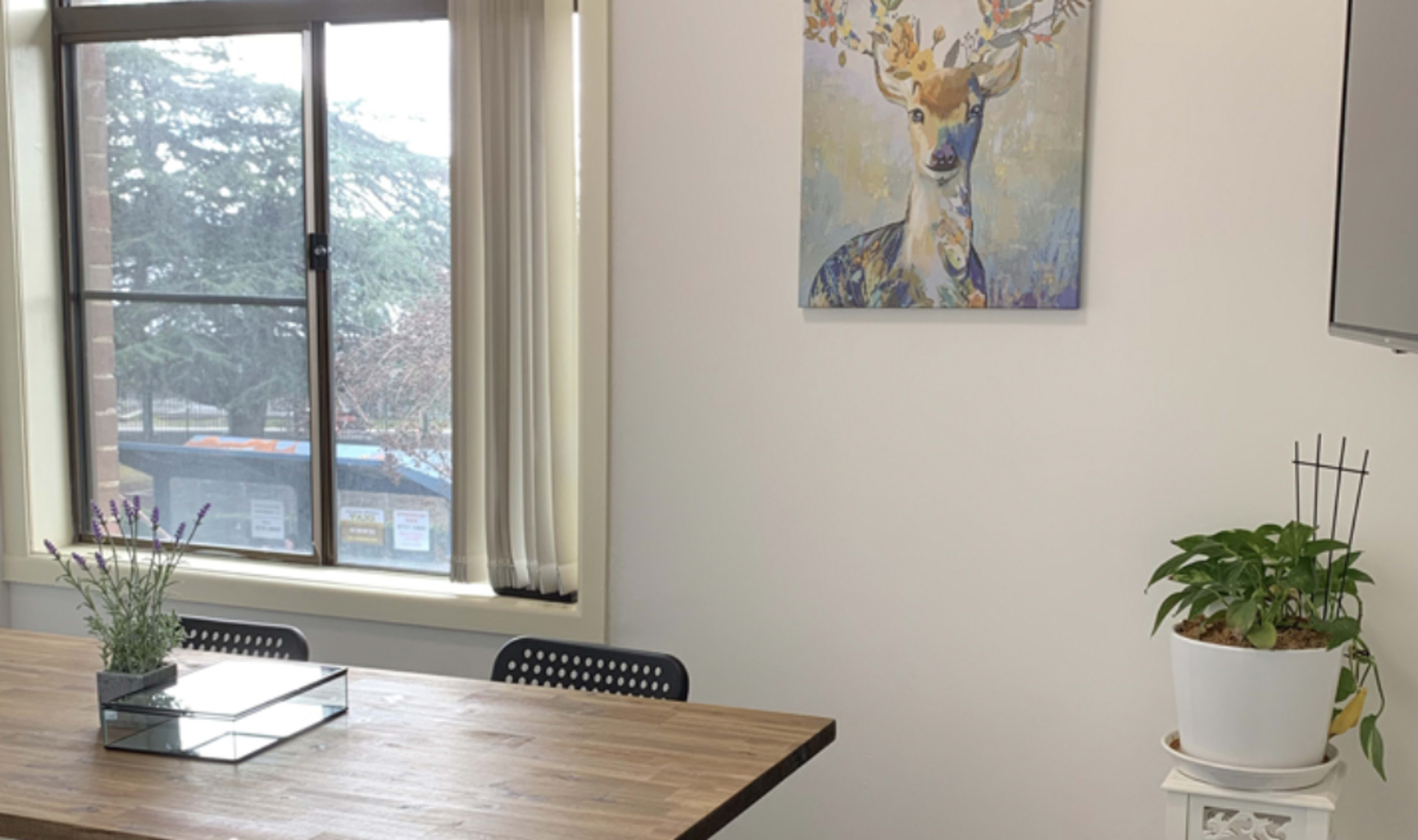 A wooden table is positioned in front of a window, with a painting of a deer on the wall and potted plants on a nearby shelf.