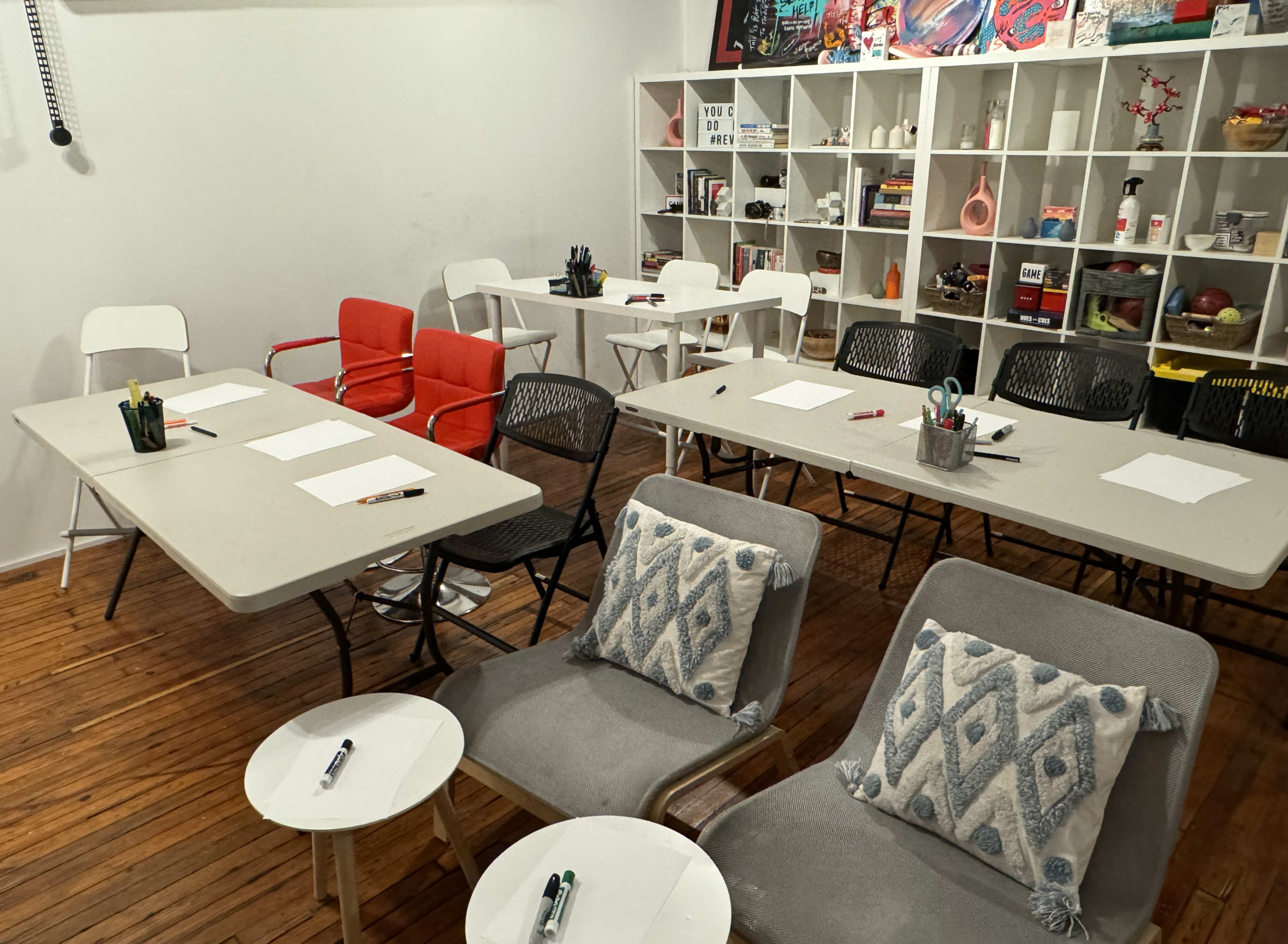 A room with several tables and chairs set up for a meeting or workshop, featuring a bookshelf filled with various items.