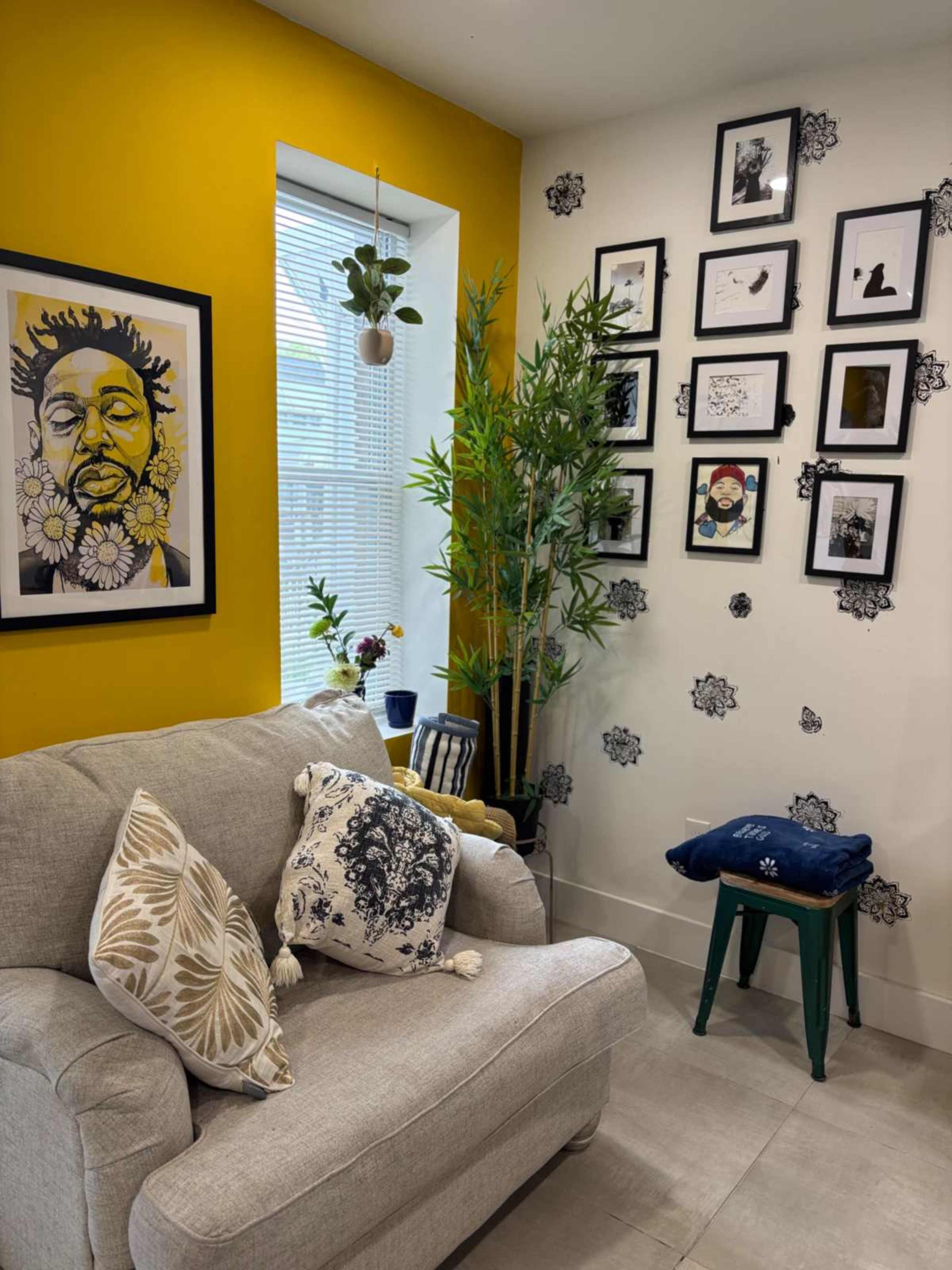 The image shows a cozy living space featuring a gray sofa adorned with decorative pillows, a large framed artwork on the yellow wall, a bamboo plant, and a gallery of framed pictures on the adjacent wall.